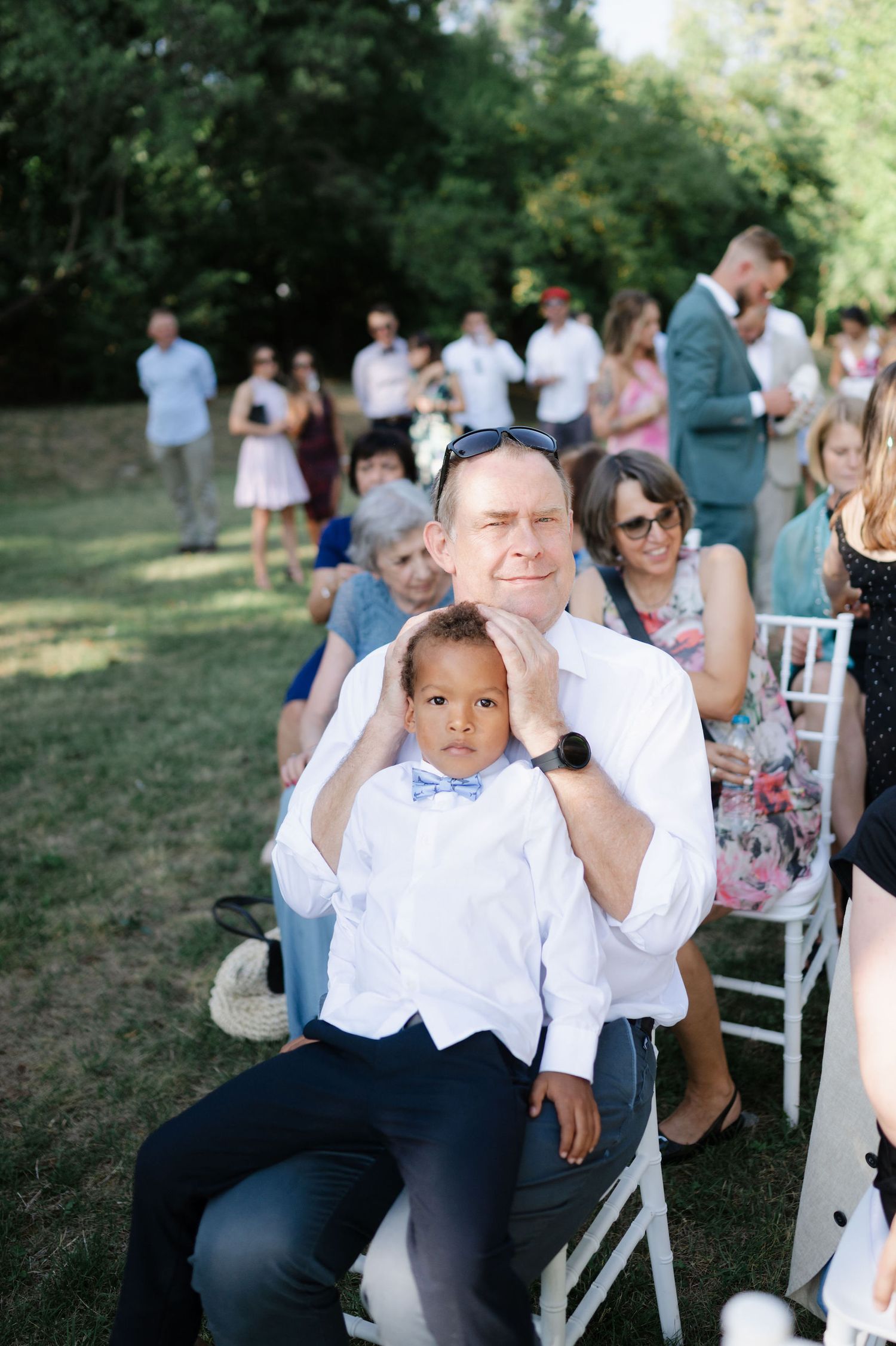 Wedding guests sit in white chairs on a grassy lawn during an outdoor ceremony.