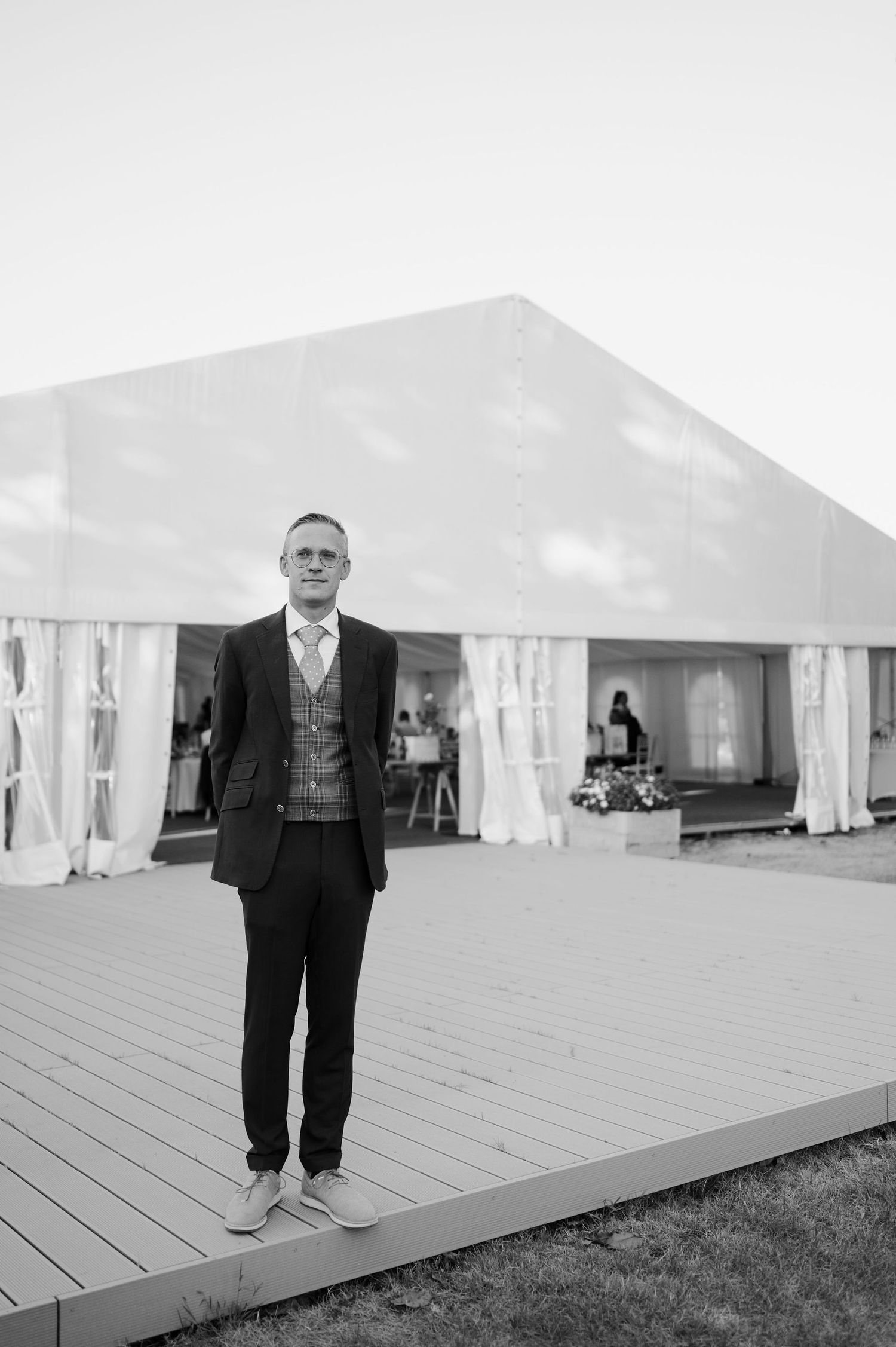 A person in formal attire standing on a wooden deck with white tents in the background.