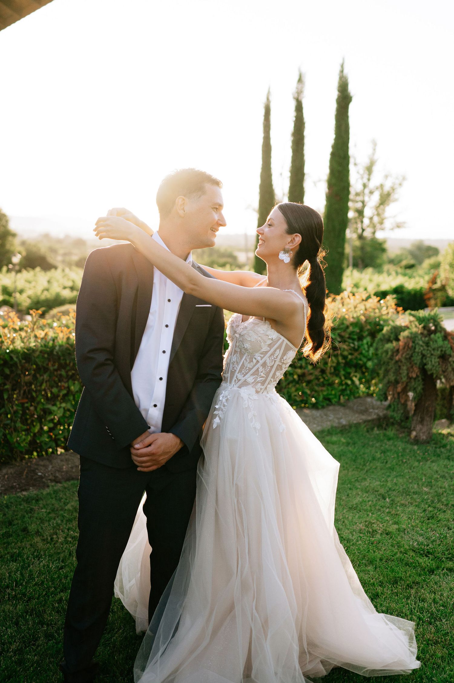 A tender moment between newlyweds dancing outdoors with sunlit cypress trees in the background.