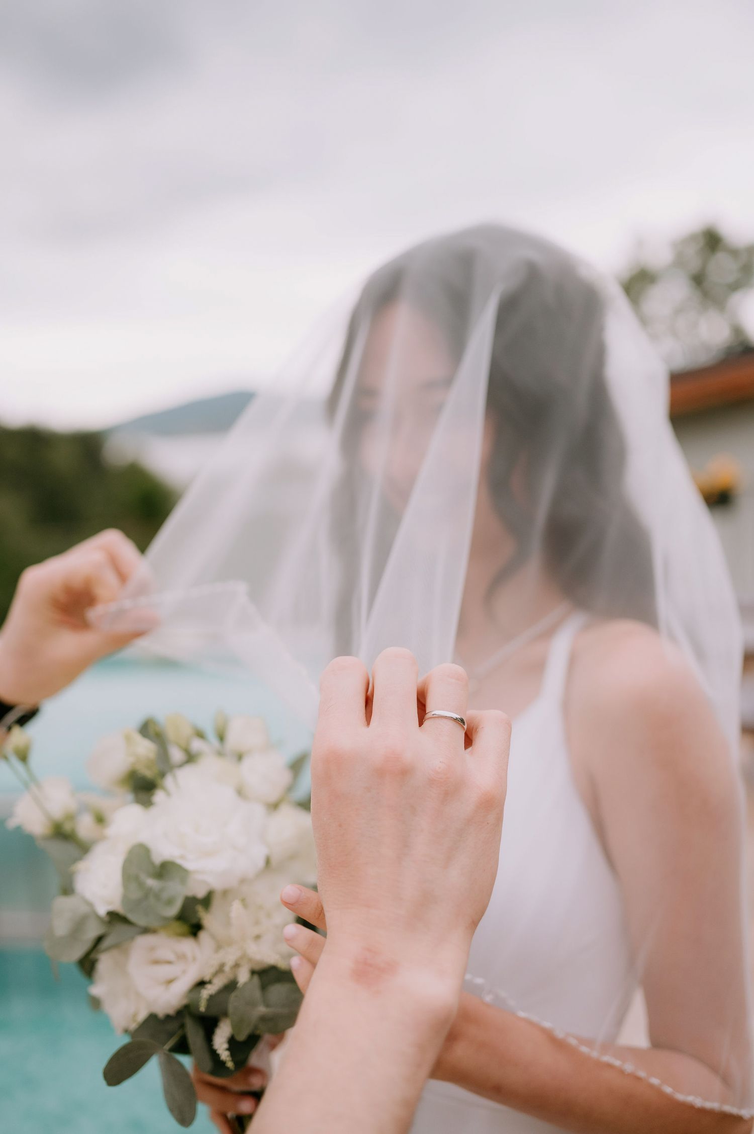 A bride wearing a white veil holds a delicate bouquet of white flowers against a blurred outdoor background.