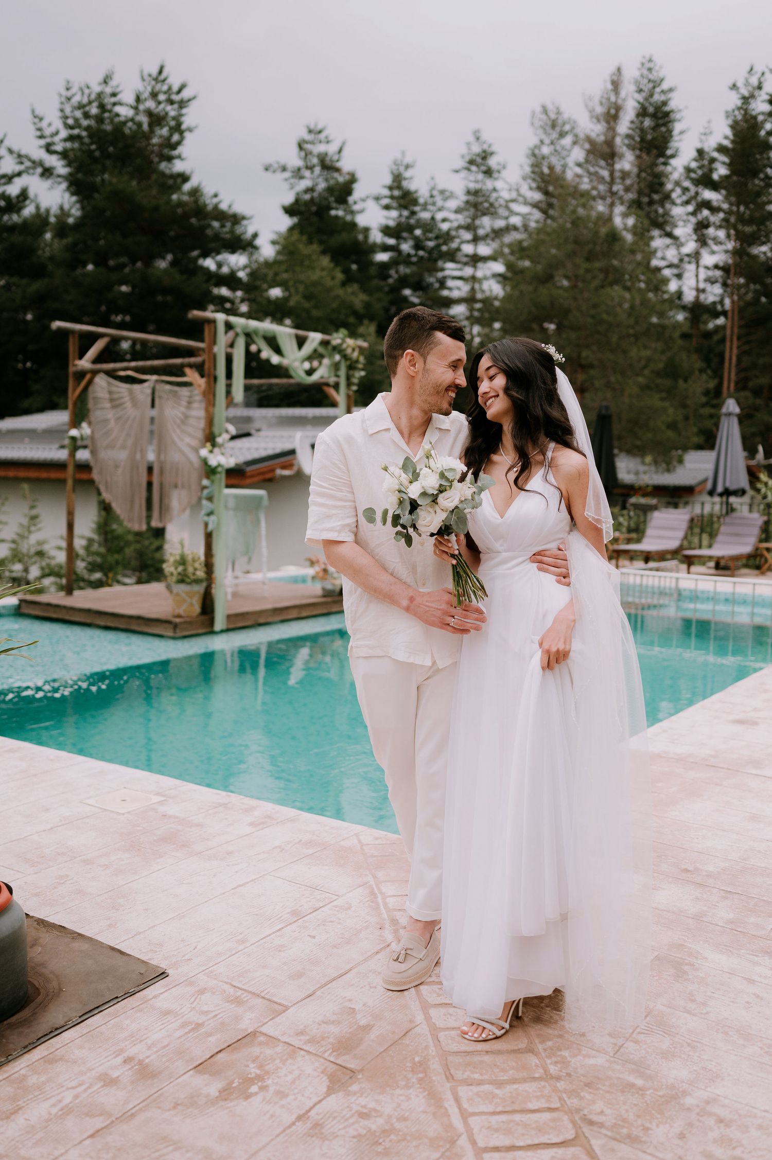Newlyweds in matching white outfits embrace near a swimming pool on their wedding day.