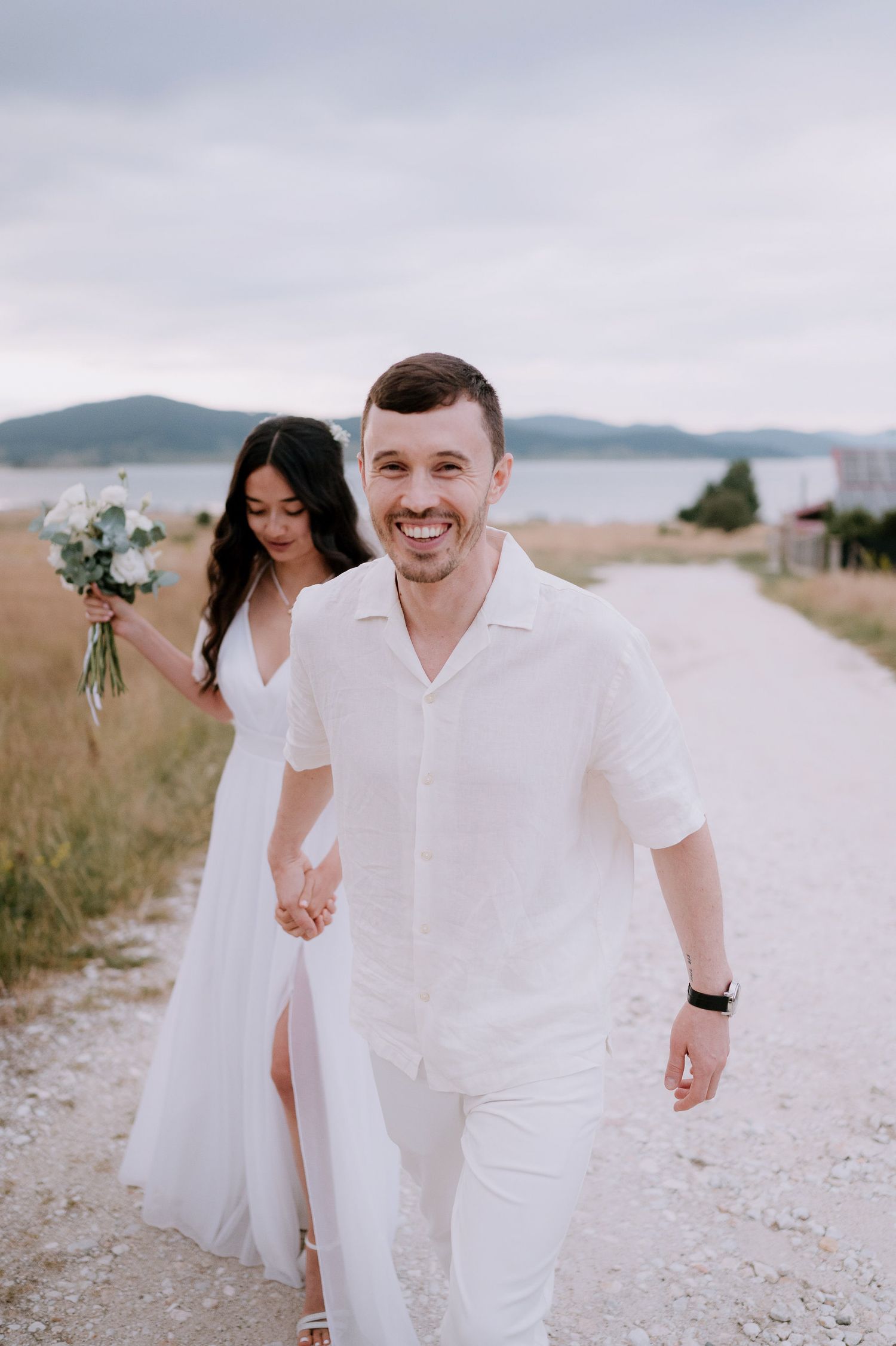 Happy couple walking along coastal path with white bouquet and flowing dress.