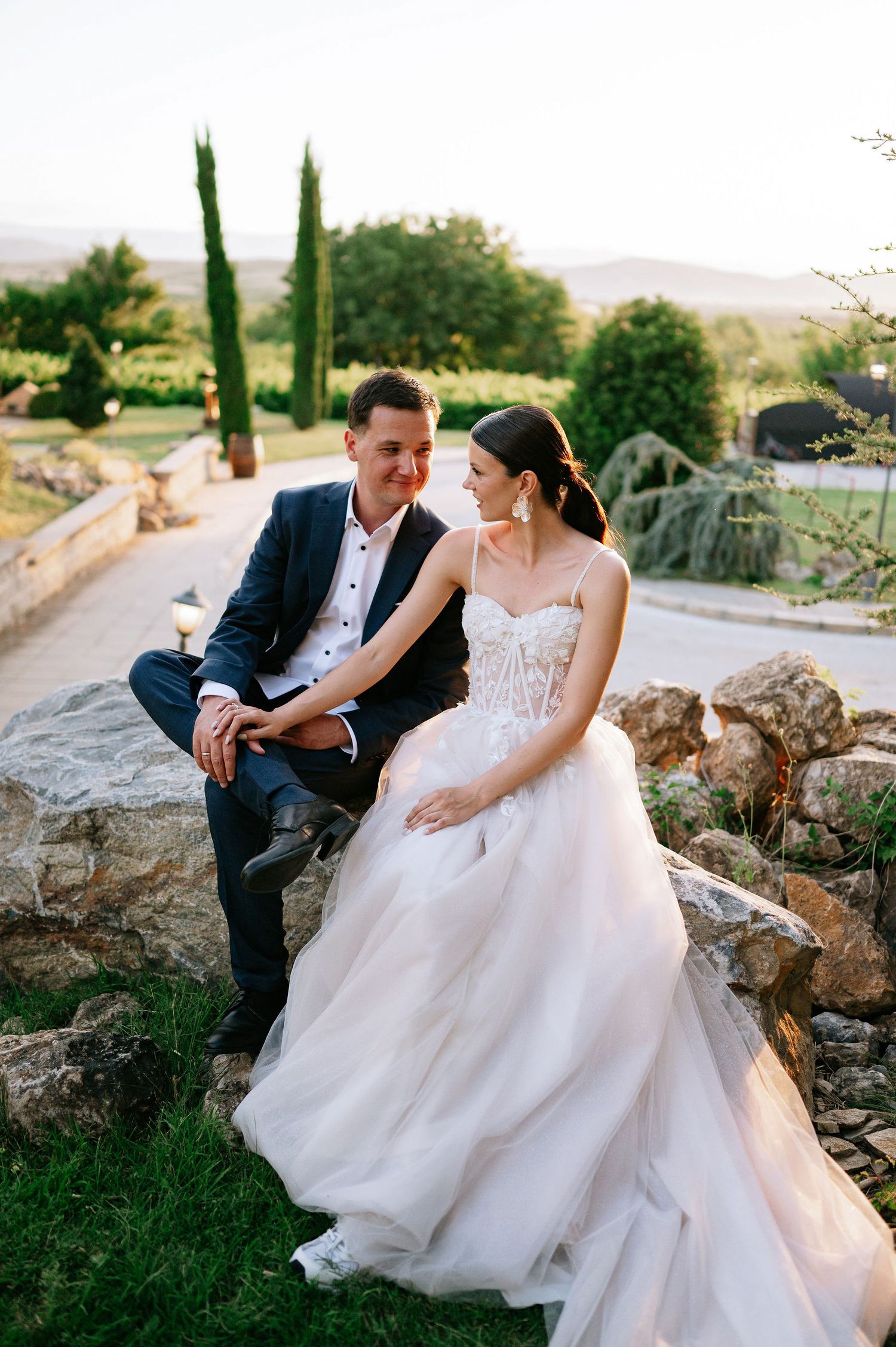 A romantic wedding couple sits together on natural stone steps surrounded by cypress trees.
