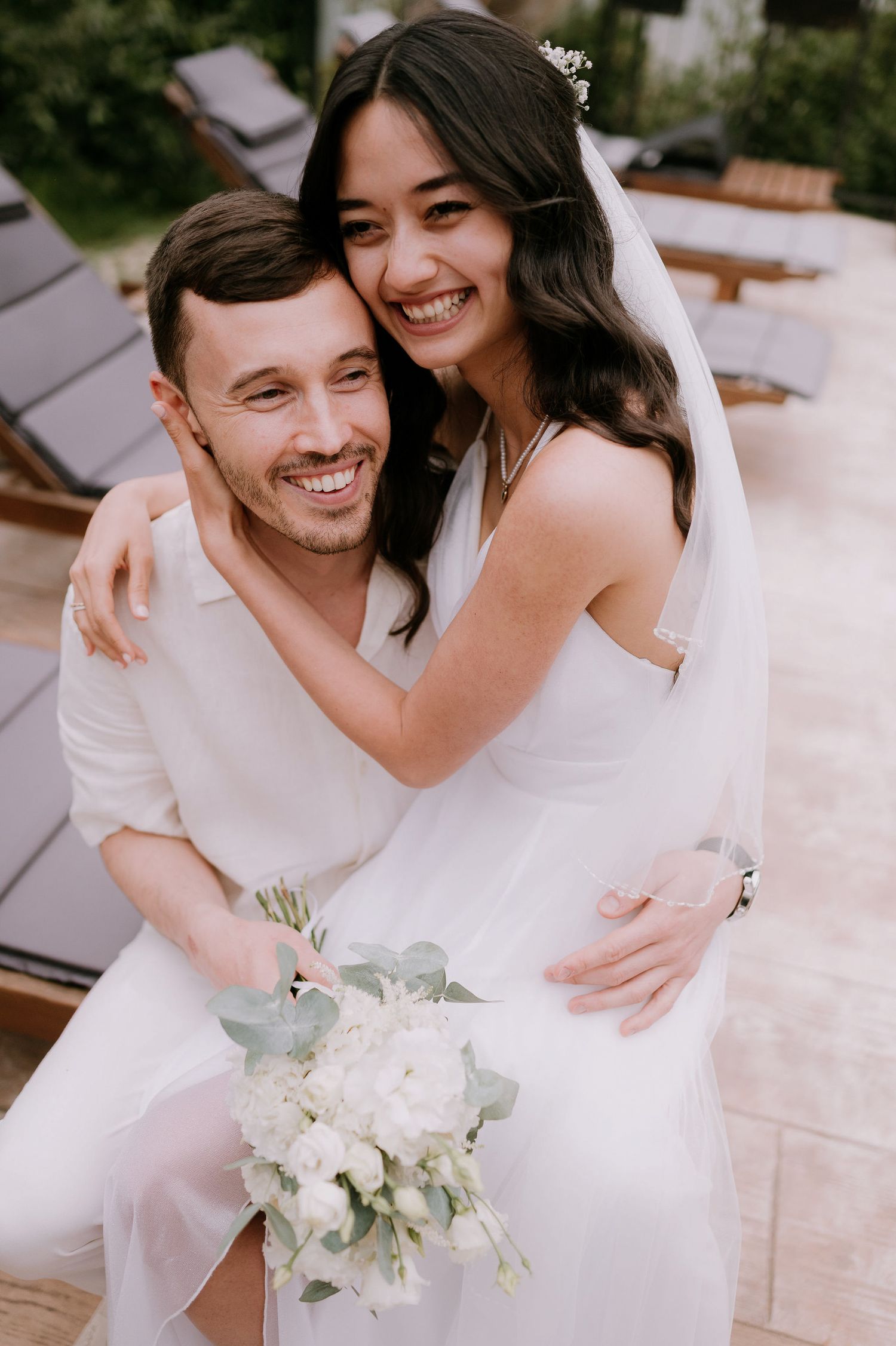 A close-up portrait of a couple in white embracing and smiling with a bridal bouquet.