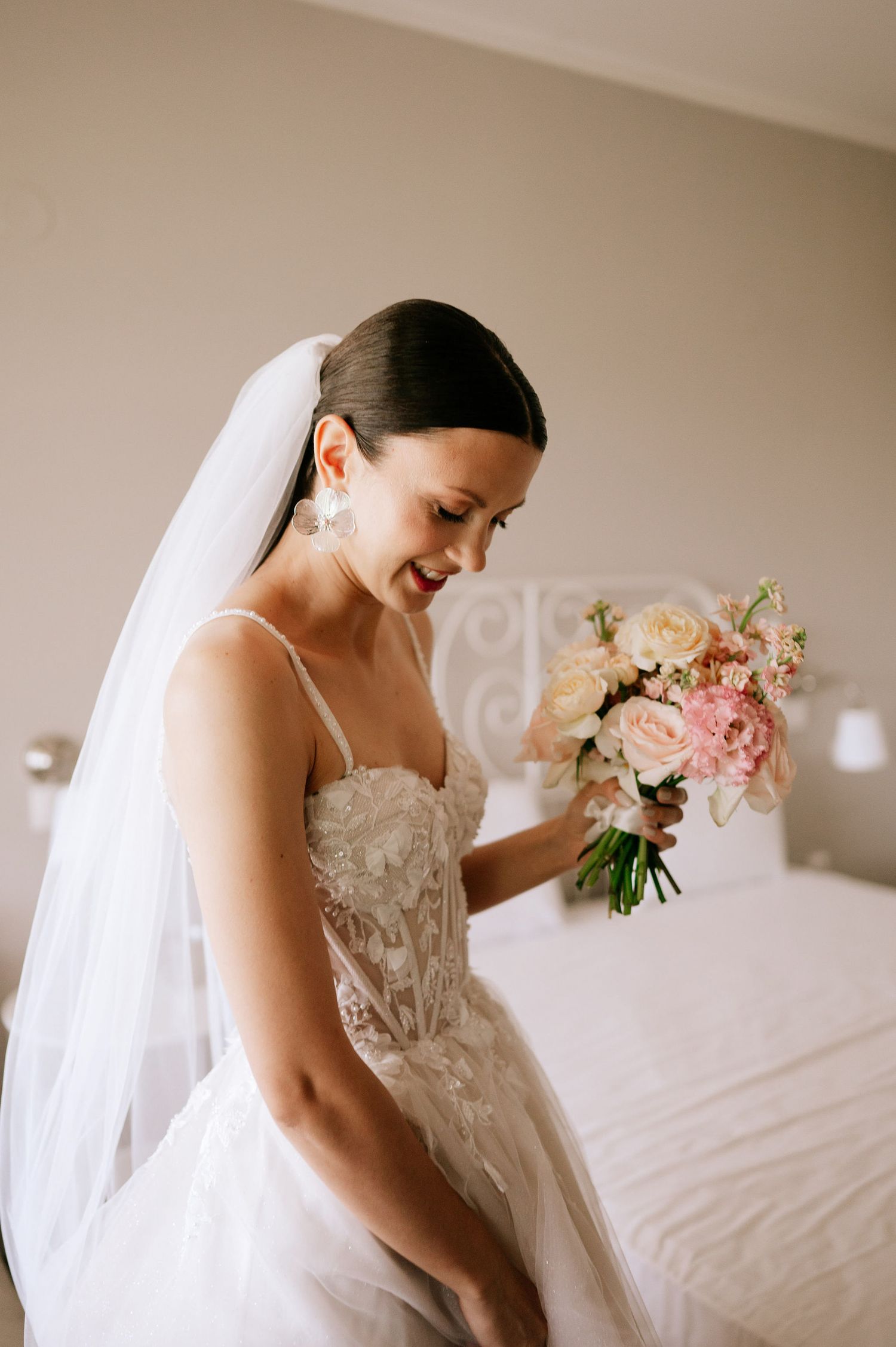 Bride in detailed white wedding dress admires her bouquet in soft natural light.