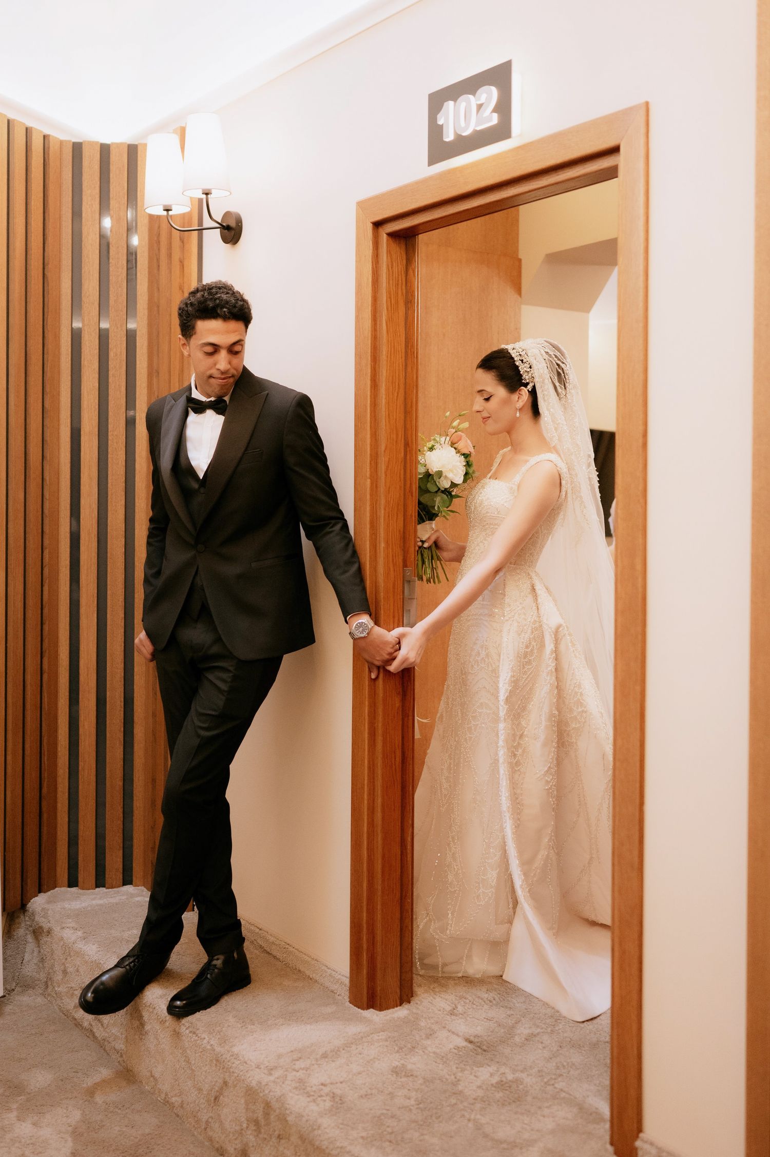 Newlyweds exit through wooden doorway holding hands in elegant wedding attire.