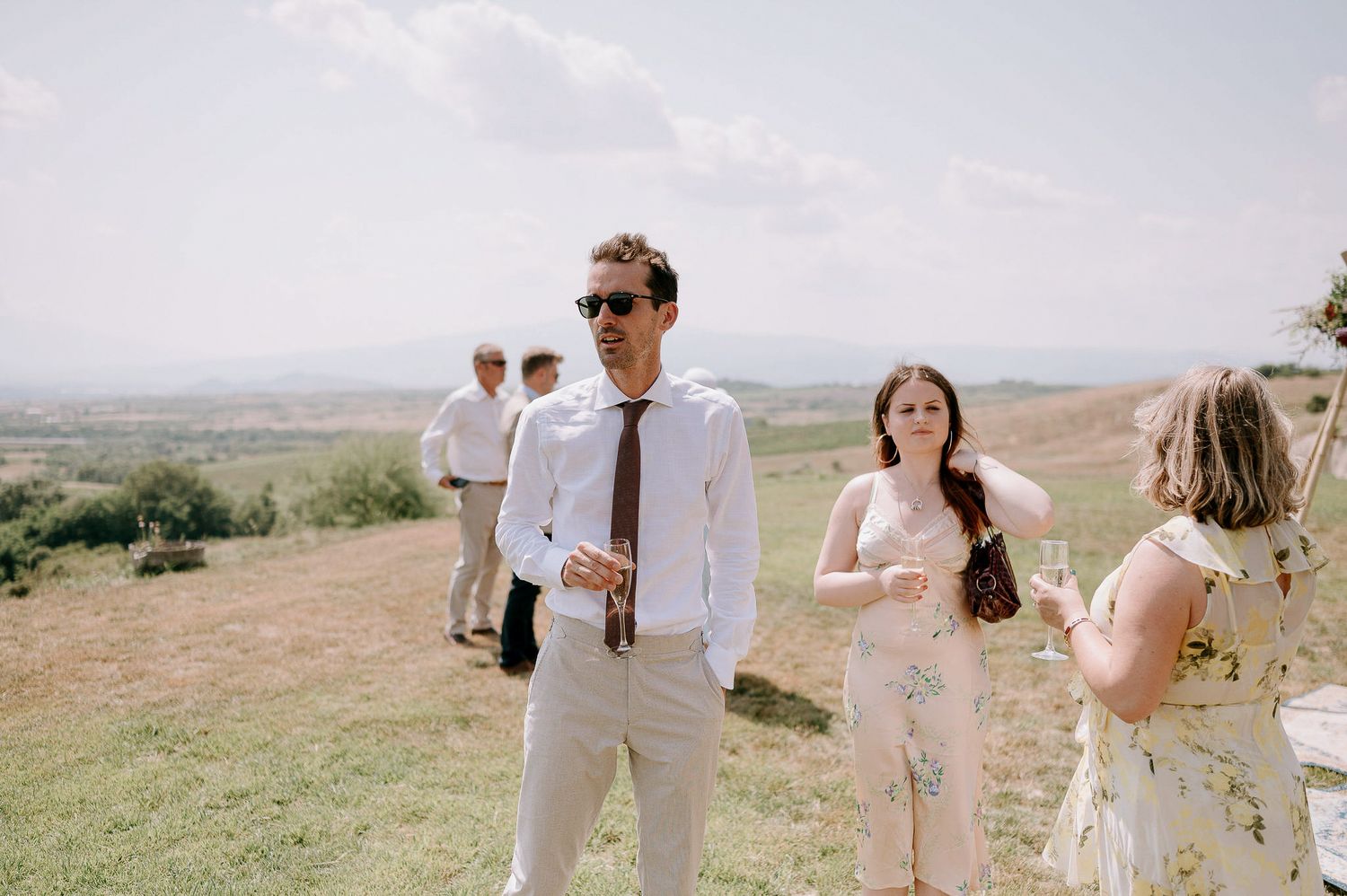 Wedding guests in light colored clothing stand on a grassy hillside with scenic views.