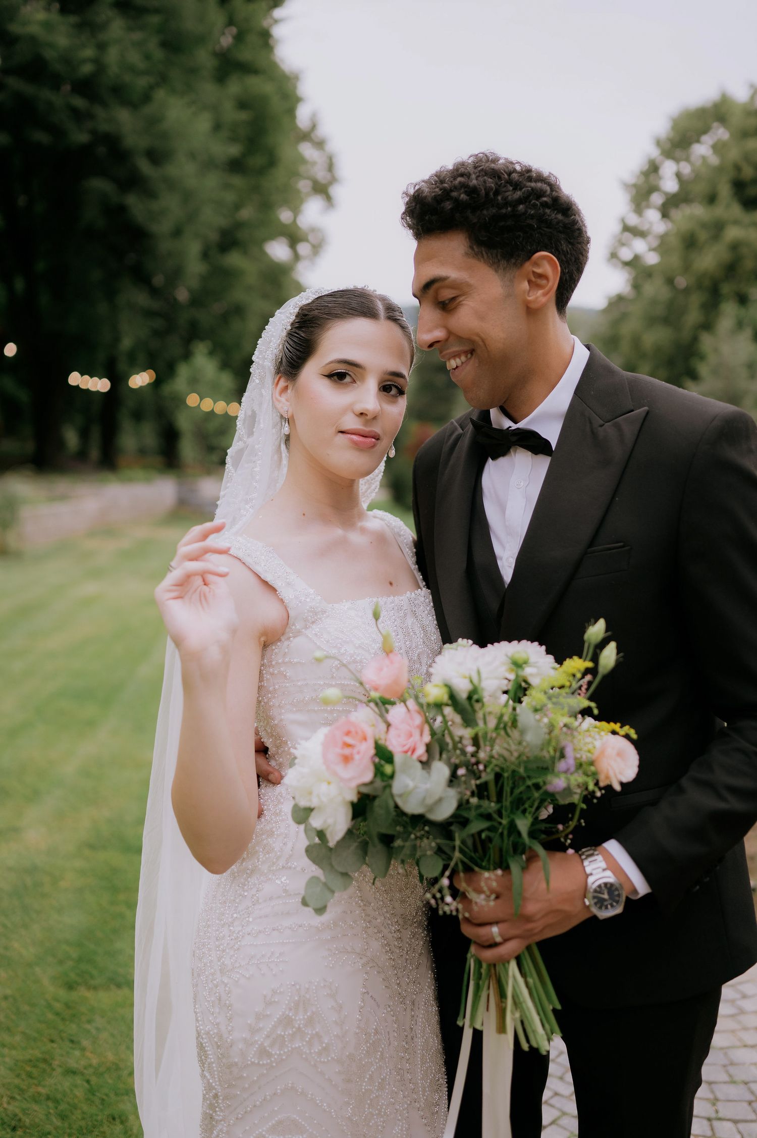 Wedding couple poses with a beautiful bouquet of white and pink flowers on a garden path.