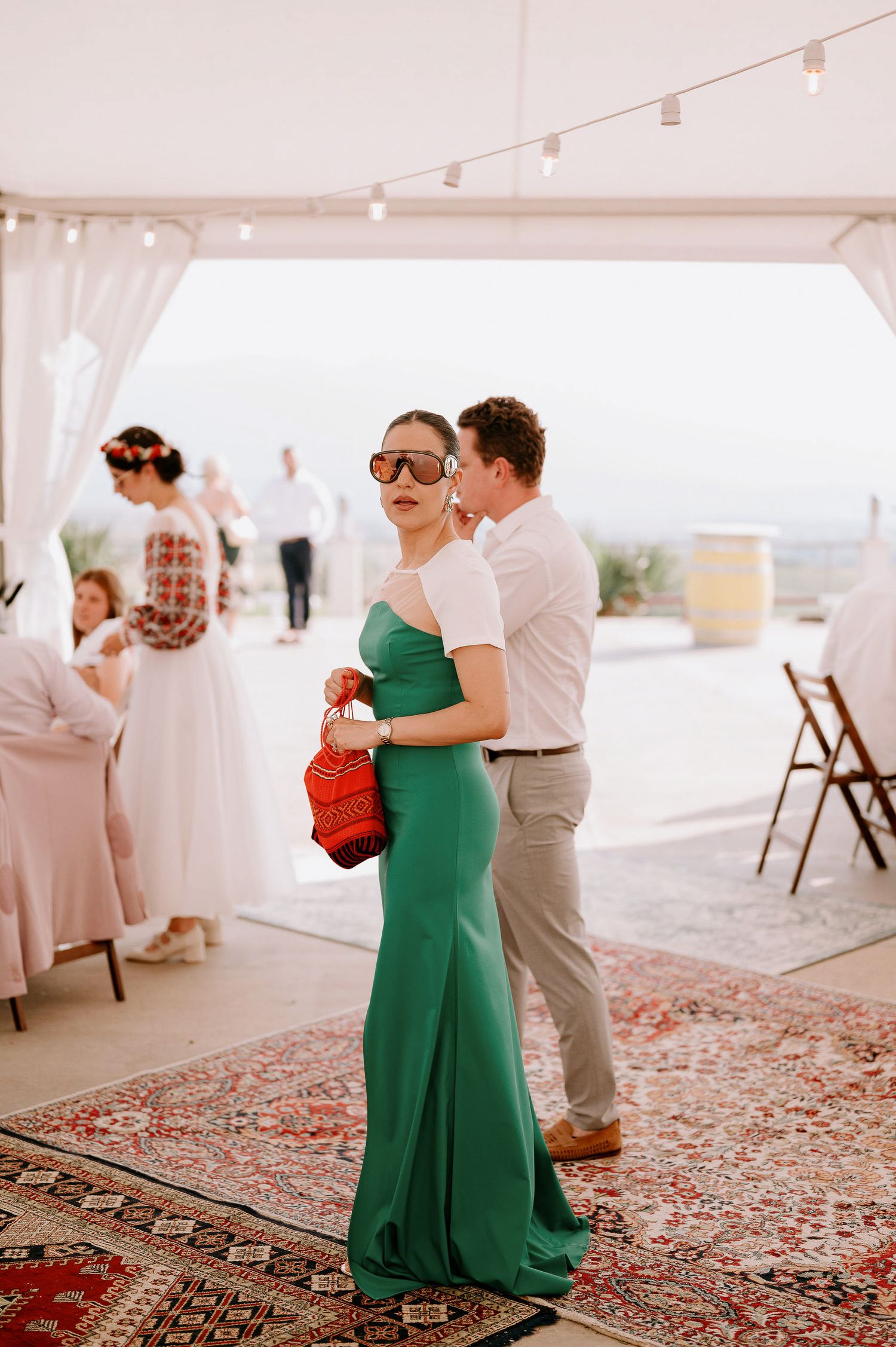 Wedding guests dancing on a patterned carpet under a white tent at an outdoor reception.