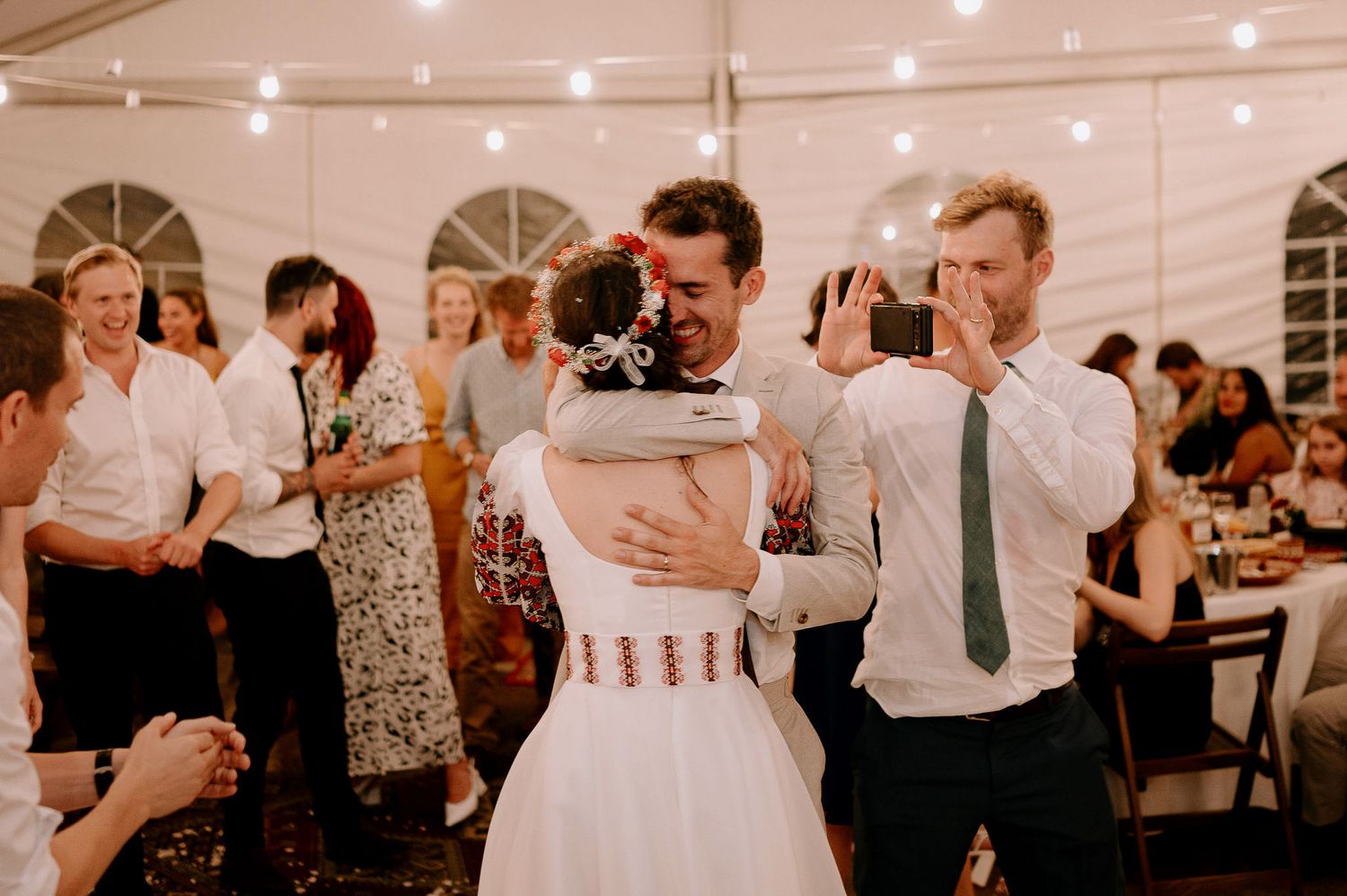 Wedding guests dancing and celebrating in a white tent with string lighting.