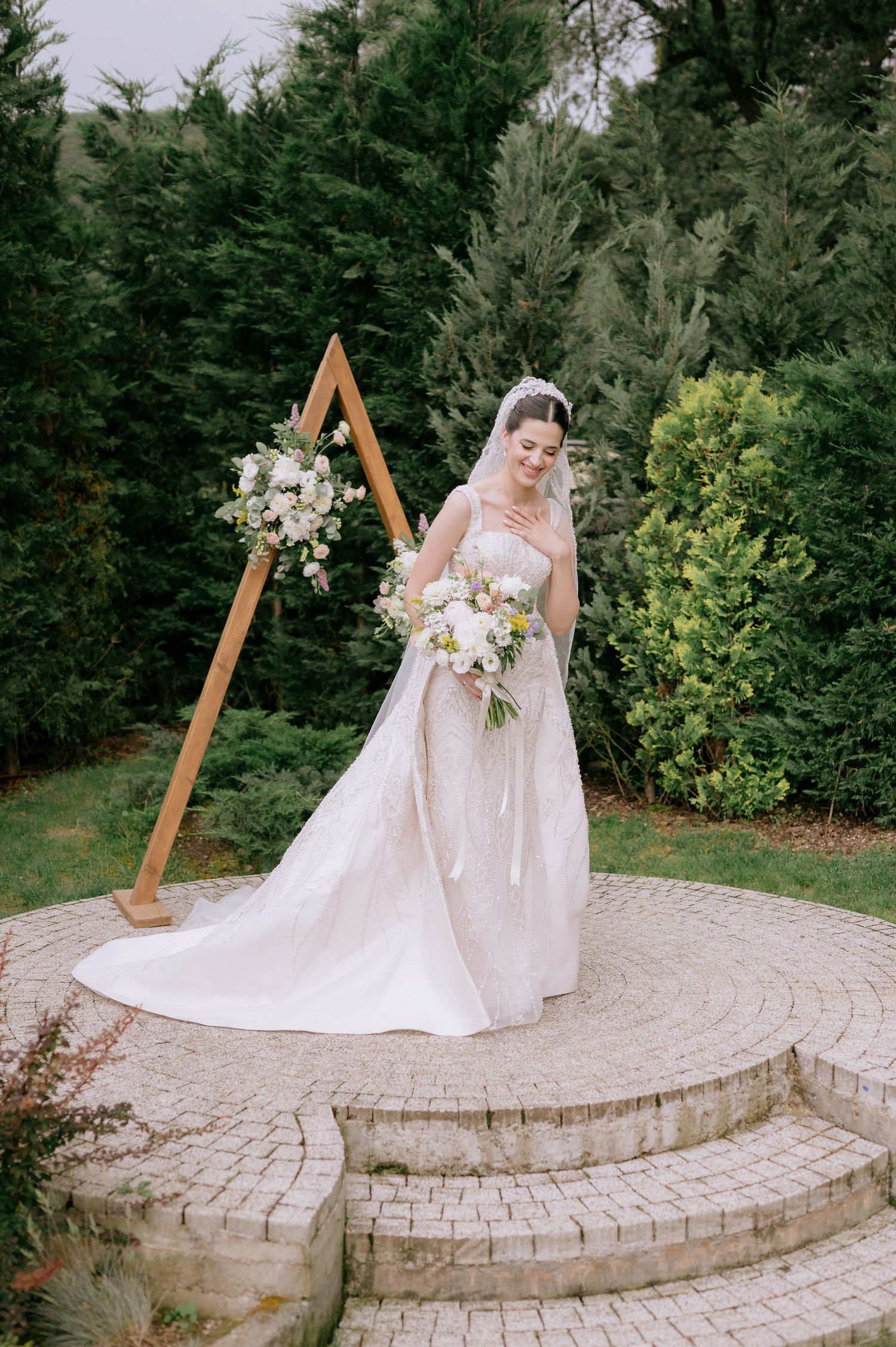 White wedding gown with flowing train displayed on curved stone patio near evergreen trees.