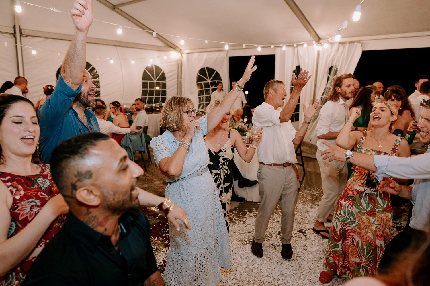 Energetic crowd dances and celebrates at a wedding reception under string lights.