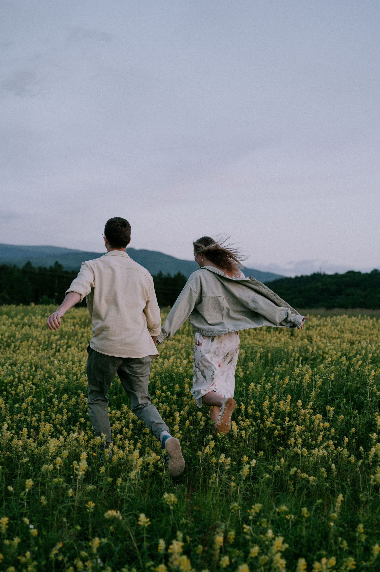 Two people run hand in hand through a vibrant yellow flower field at dusk.