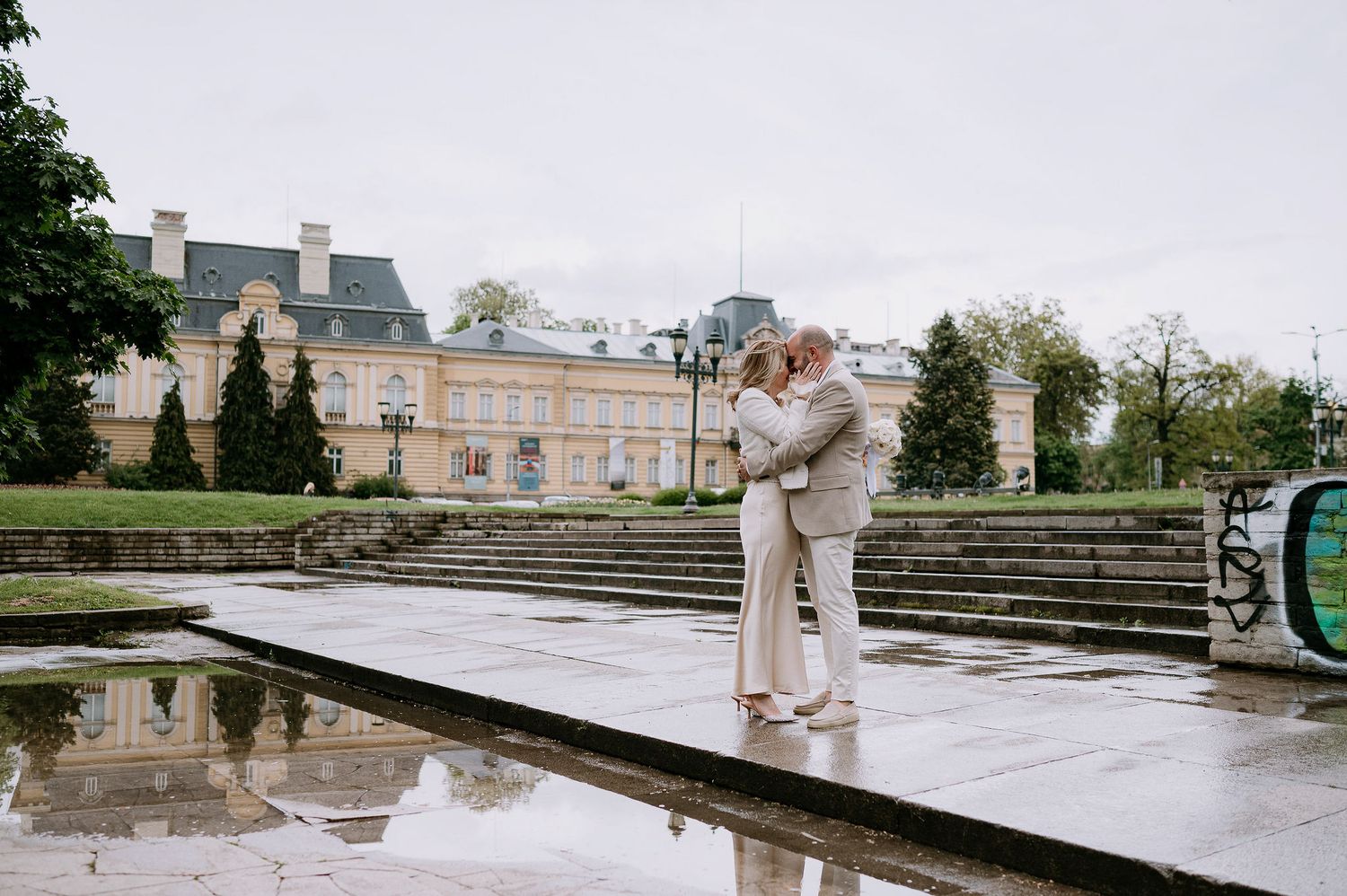 A couple in light colored outfits poses near stone steps and a reflecting pool at an elegant estate.