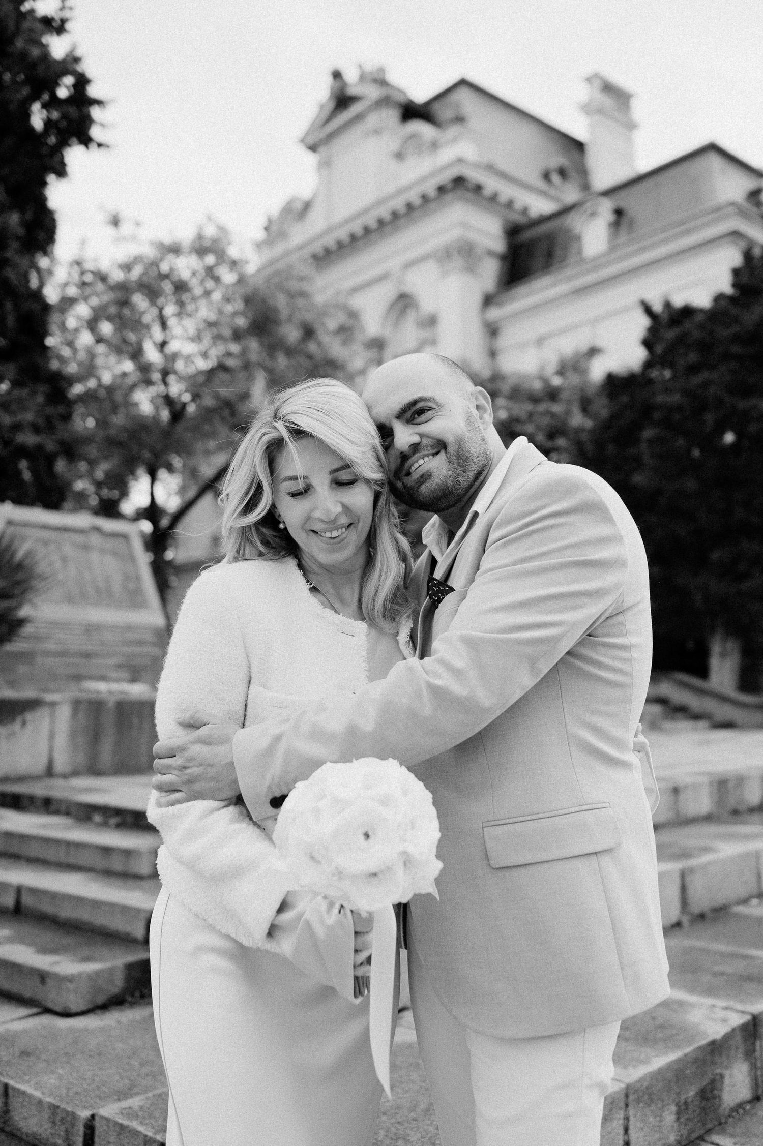 A joyful couple in white shares laughter and embraces near a historic building.