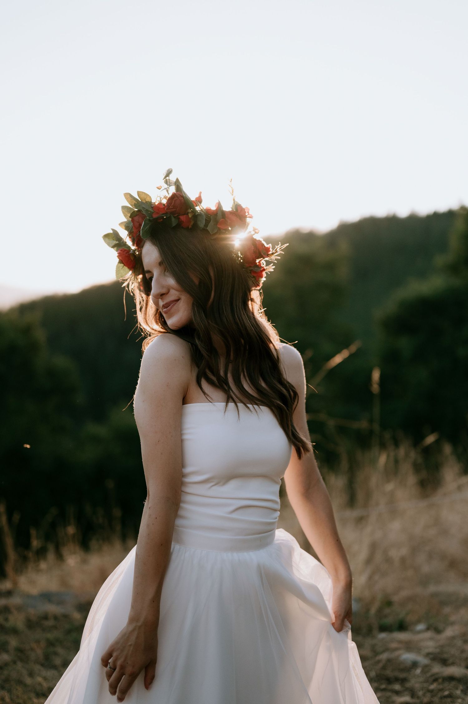 A flowing white wedding dress and floral crown silhouetted against a golden sunset in a mountain field.