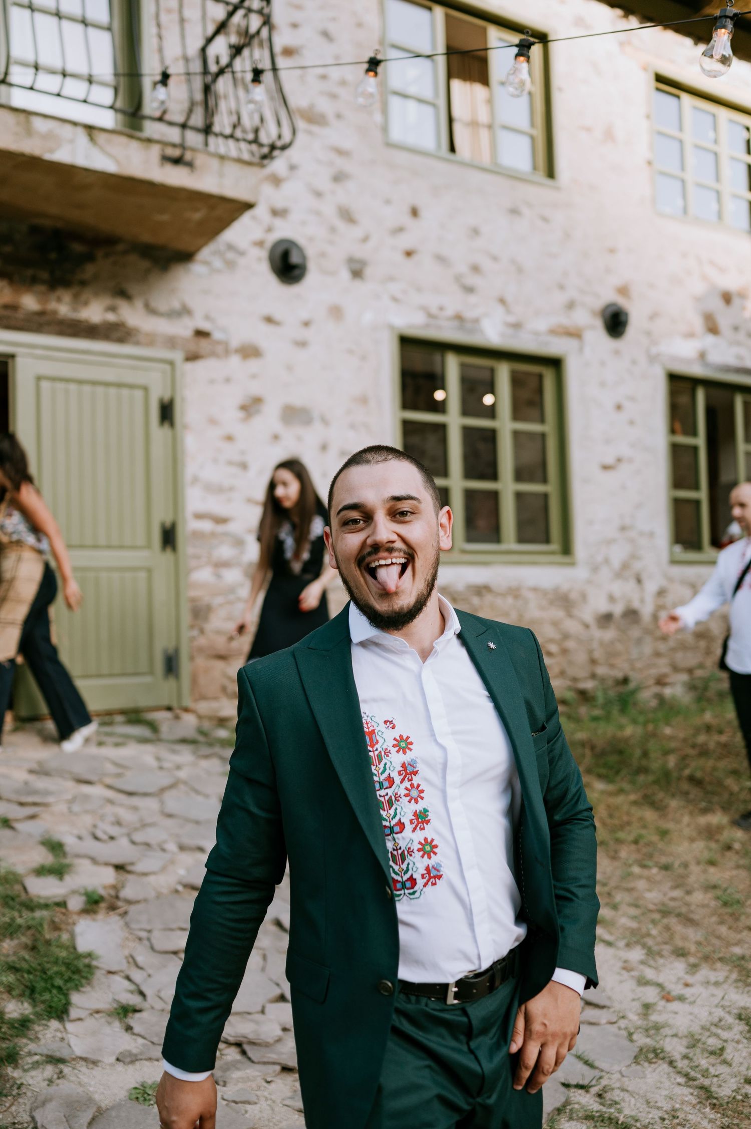 Person in green suit with embroidered shirt posing joyfully outside stone building.