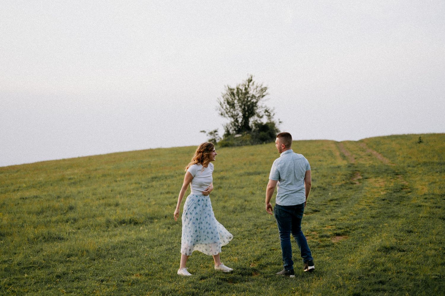 Two people walk together across a scenic green meadow with mountains in the distance.