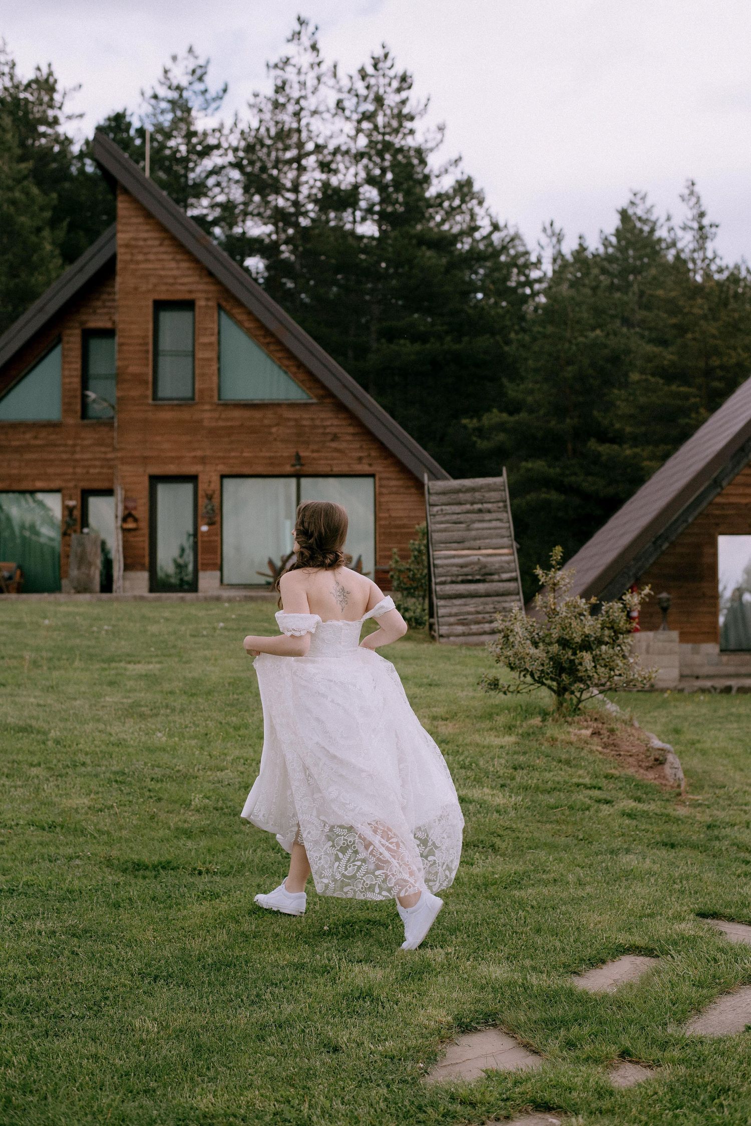 A white dress flows in the breeze as figures walk near rustic cabins on a grassy hill.