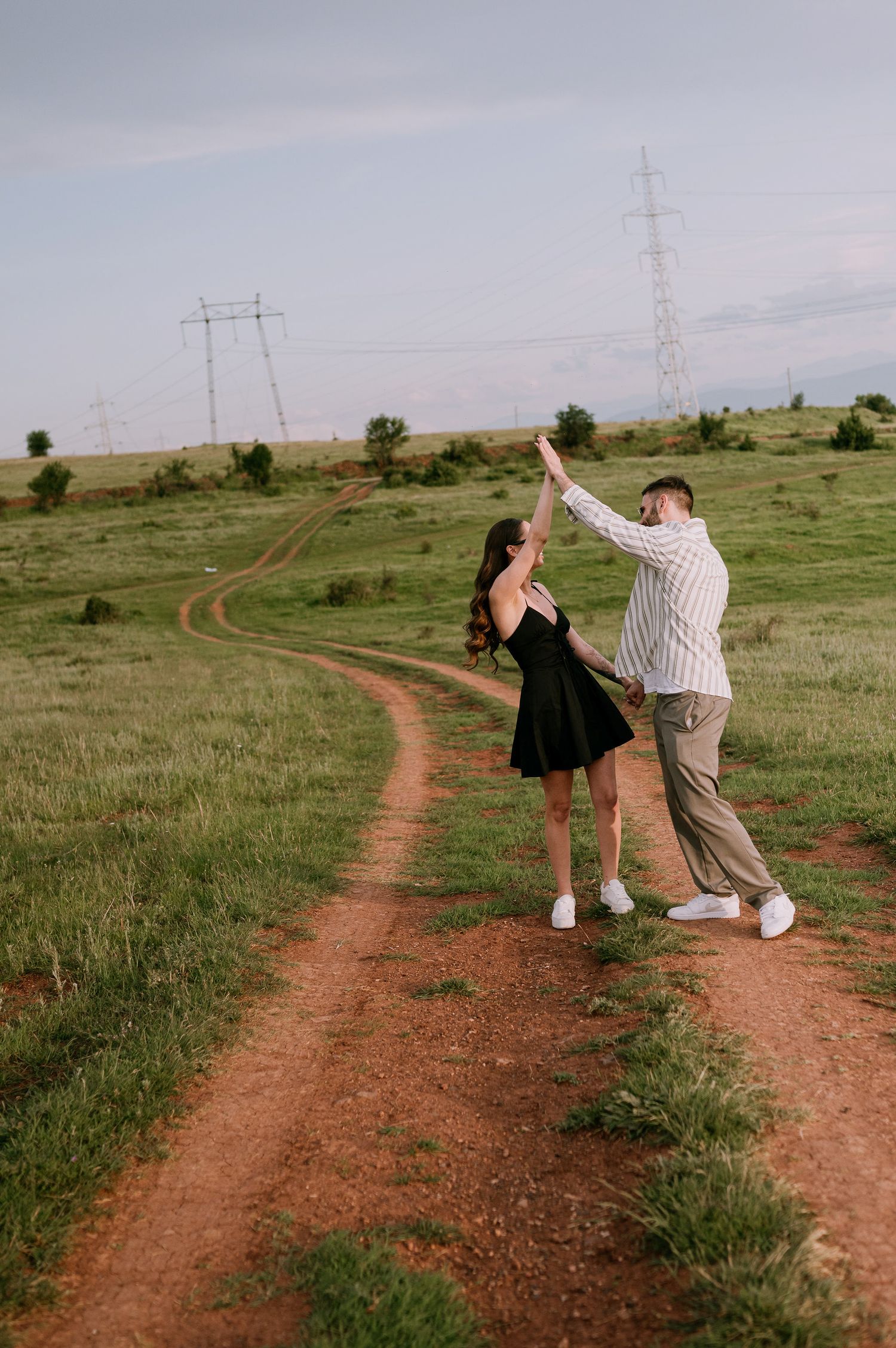 Two people dance playfully together on a winding dirt path through green grassland with power lines in the distance.