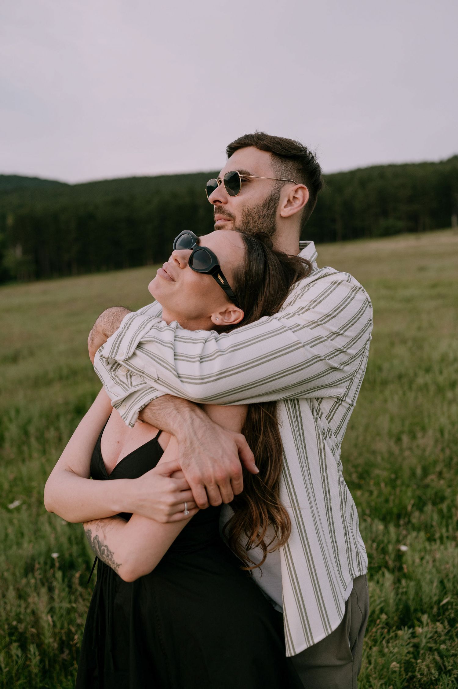 A couple embraces intimately in a grassy field during sunset, wearing casual attire and sunglasses.
