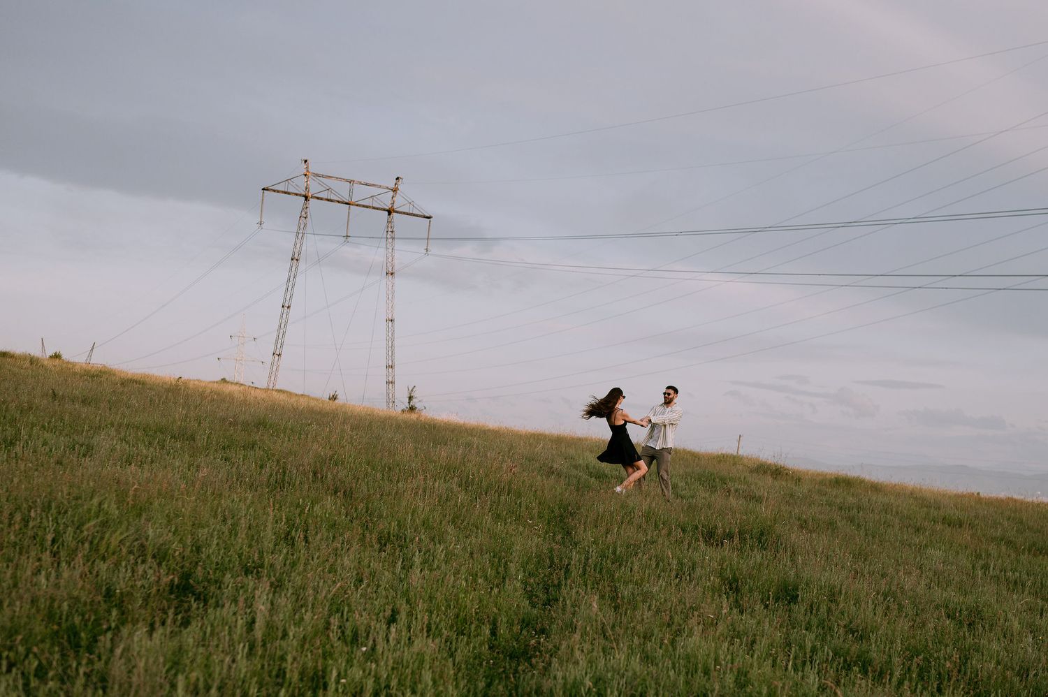 A person walks through a grassy field with power lines and utility poles stretching into the distance.