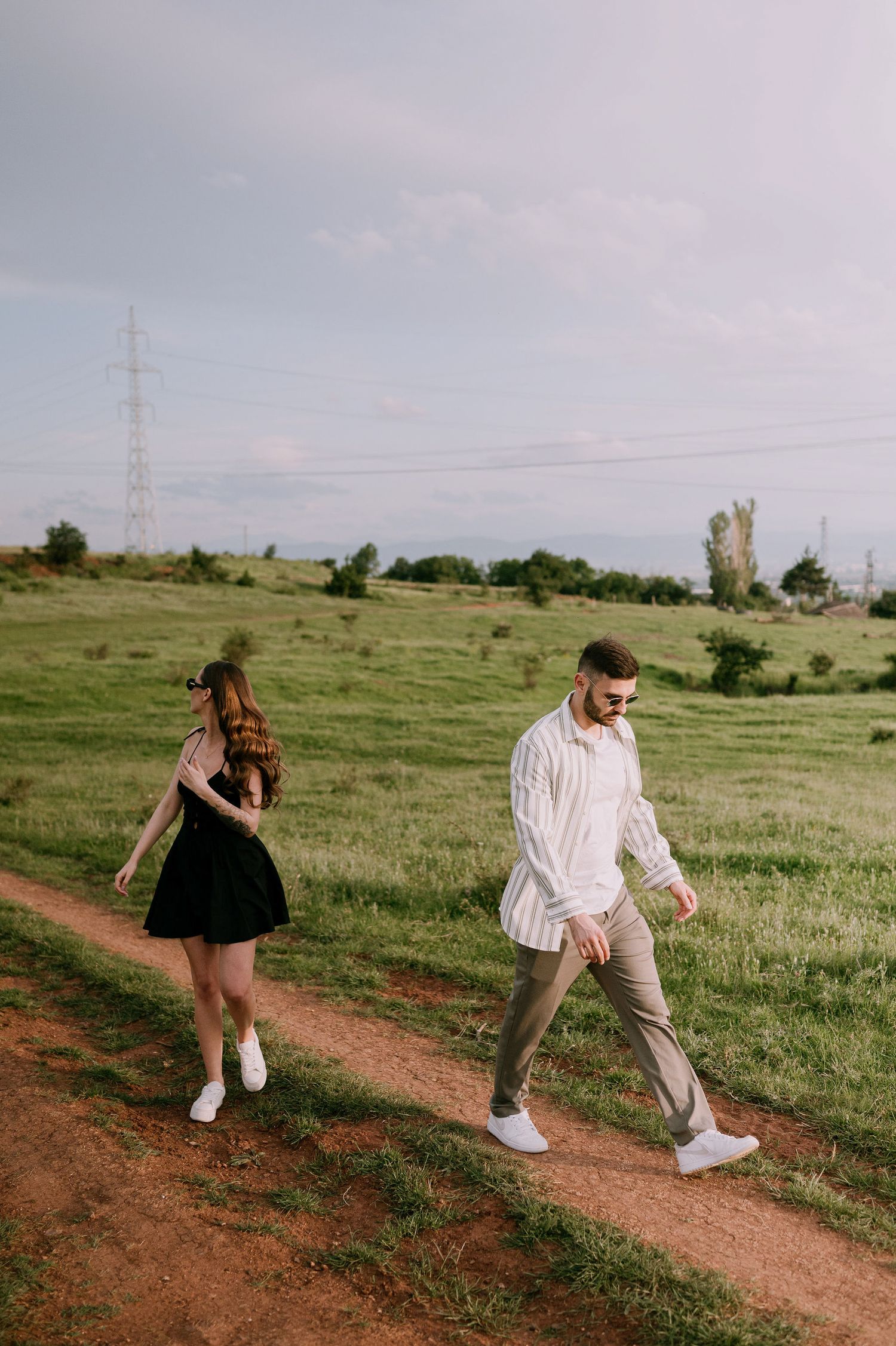 A playful couple runs and dances along a dirt path through a green meadow with power lines in the distance.