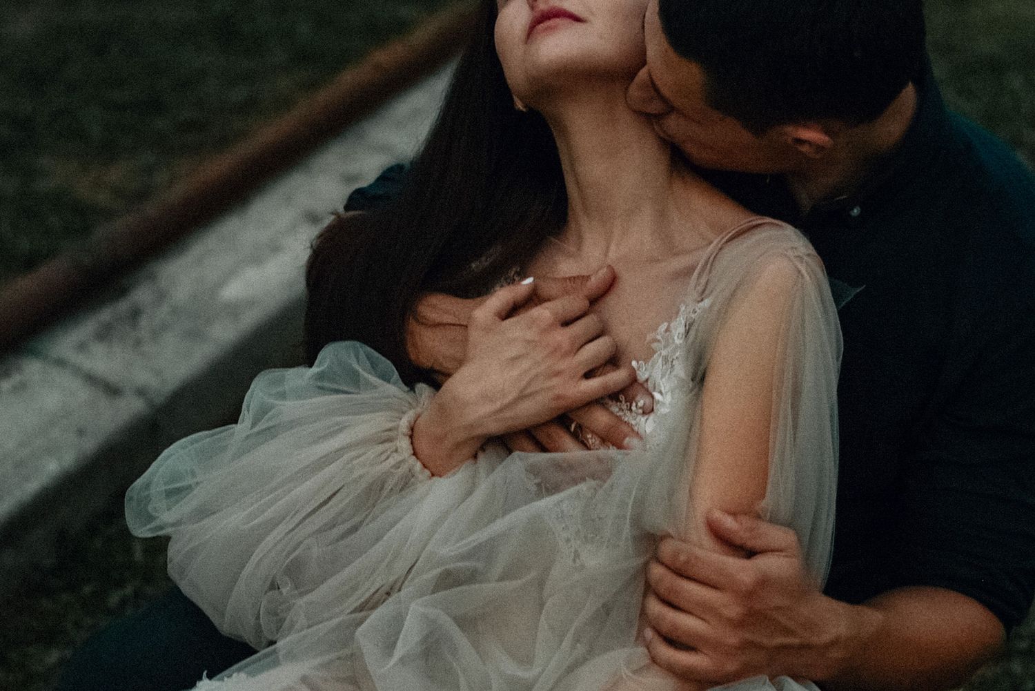 A romantic wedding couple embraces in a moody outdoor setting with soft lighting and flowing white dress details.