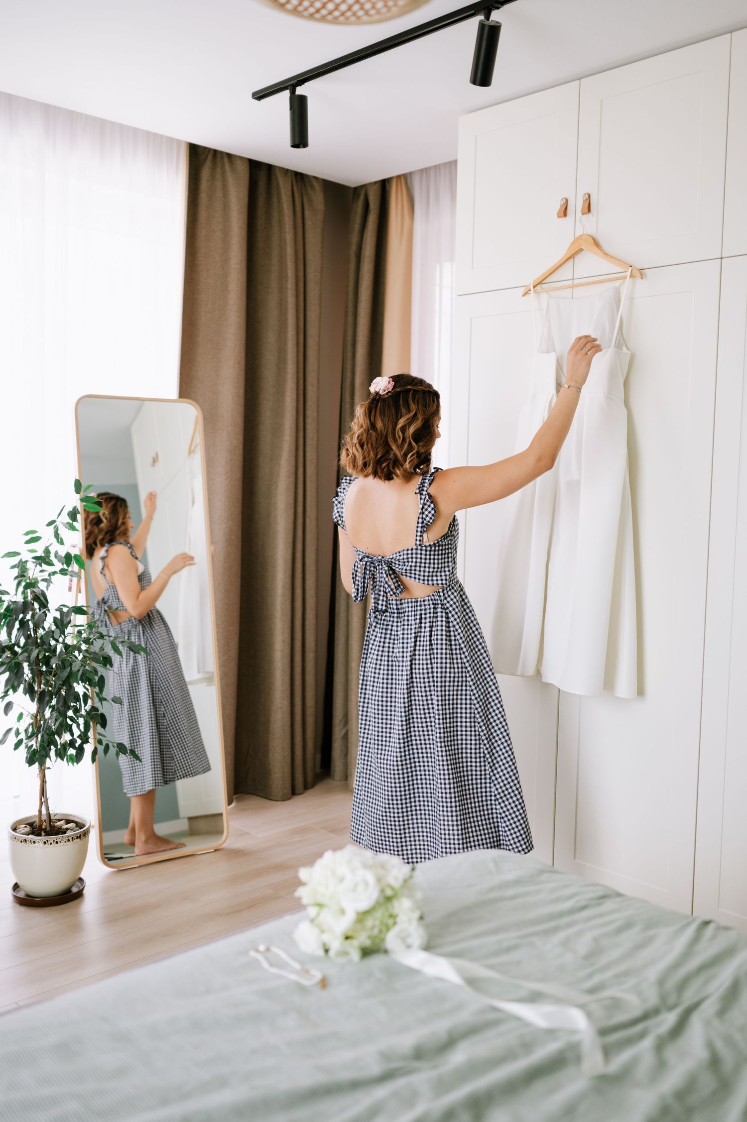 A person in a gingham dress hangs clothes on a wall-mounted rack in a bright, minimalist bedroom with houseplants.