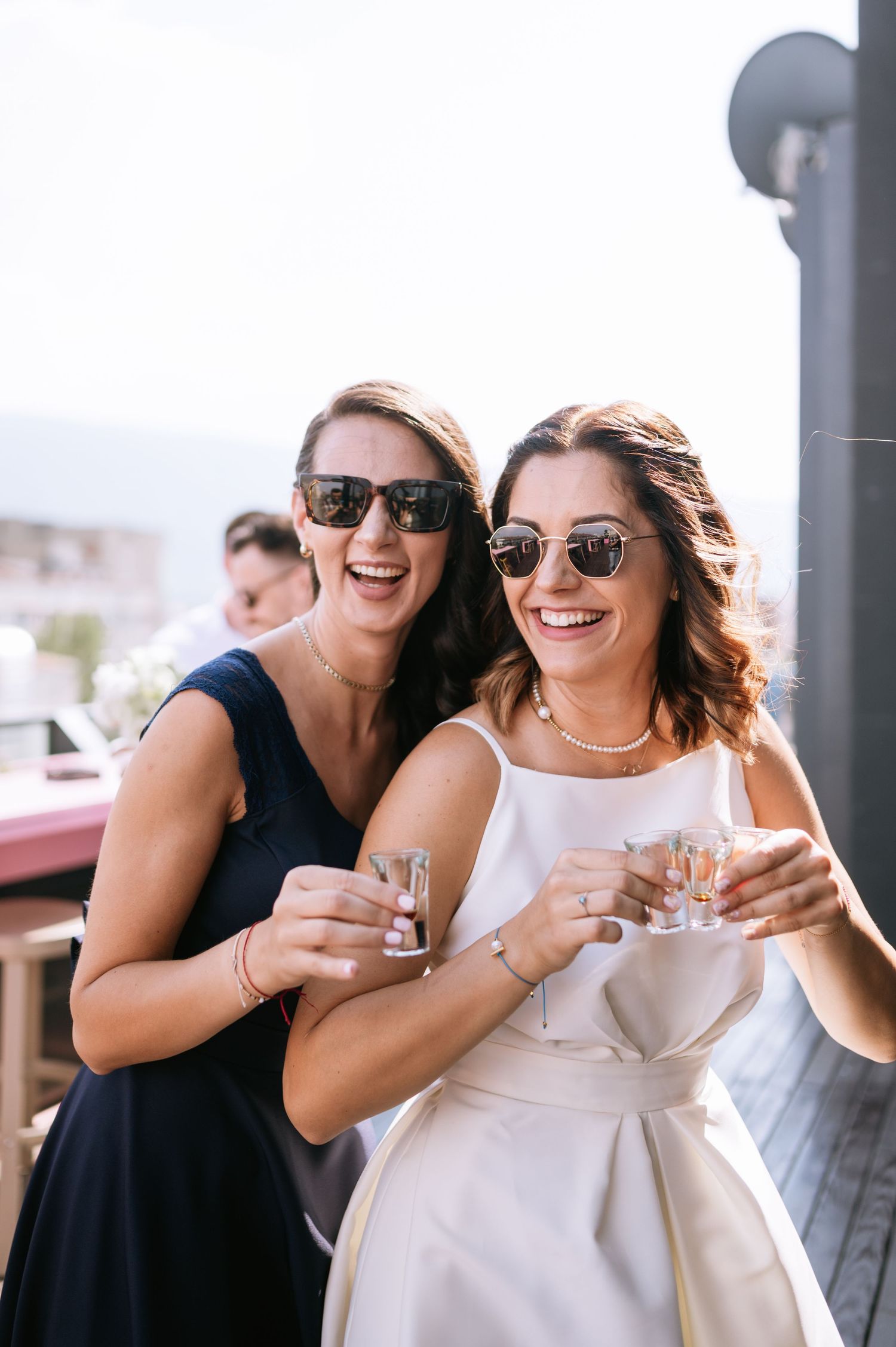 Two friends in sunglasses share drinks and laughter at an outdoor celebration, one wearing black and one in white.