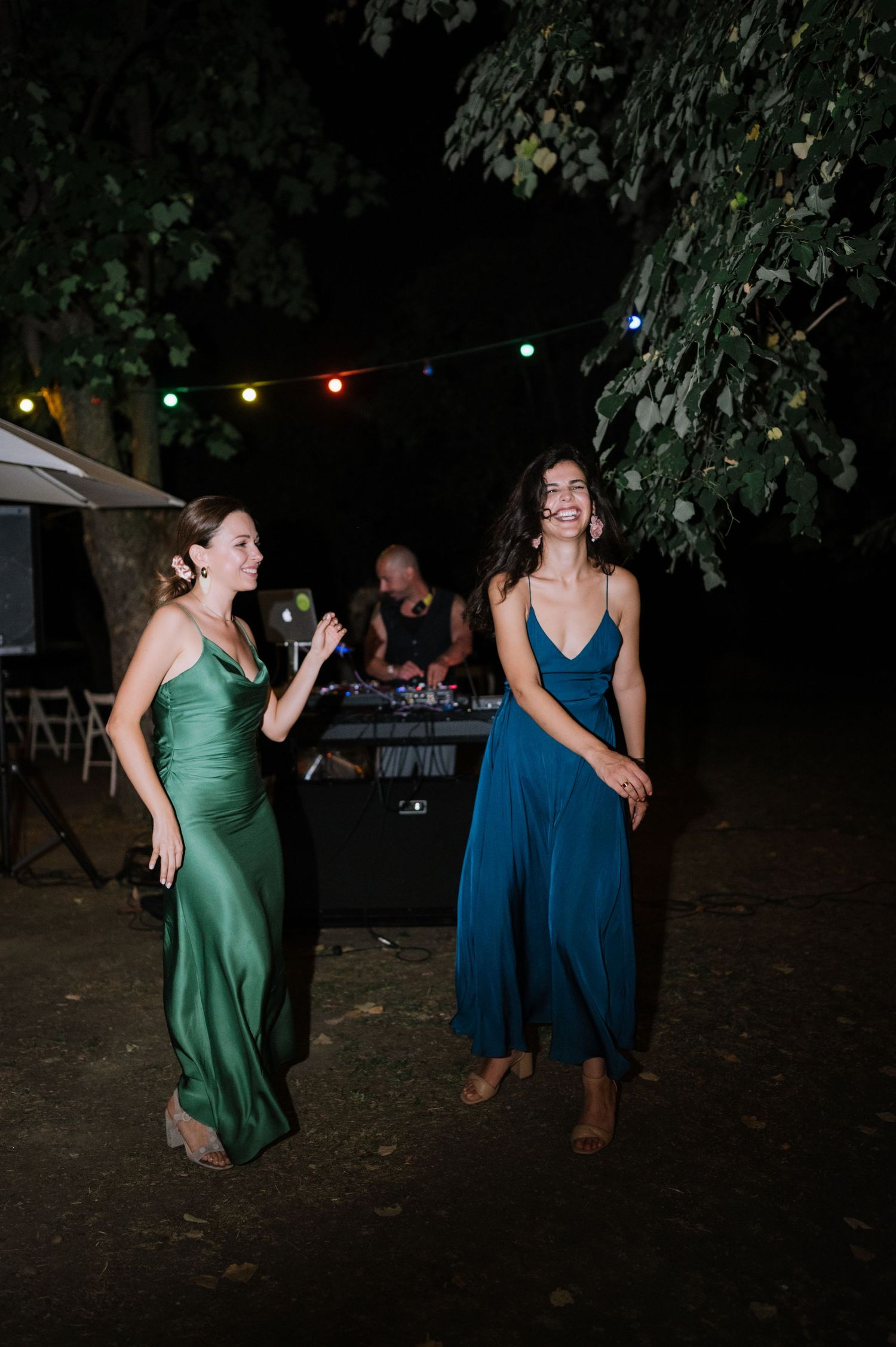 Two women in contrasting blue and green dresses dance together at an outdoor nighttime celebration under string lights.