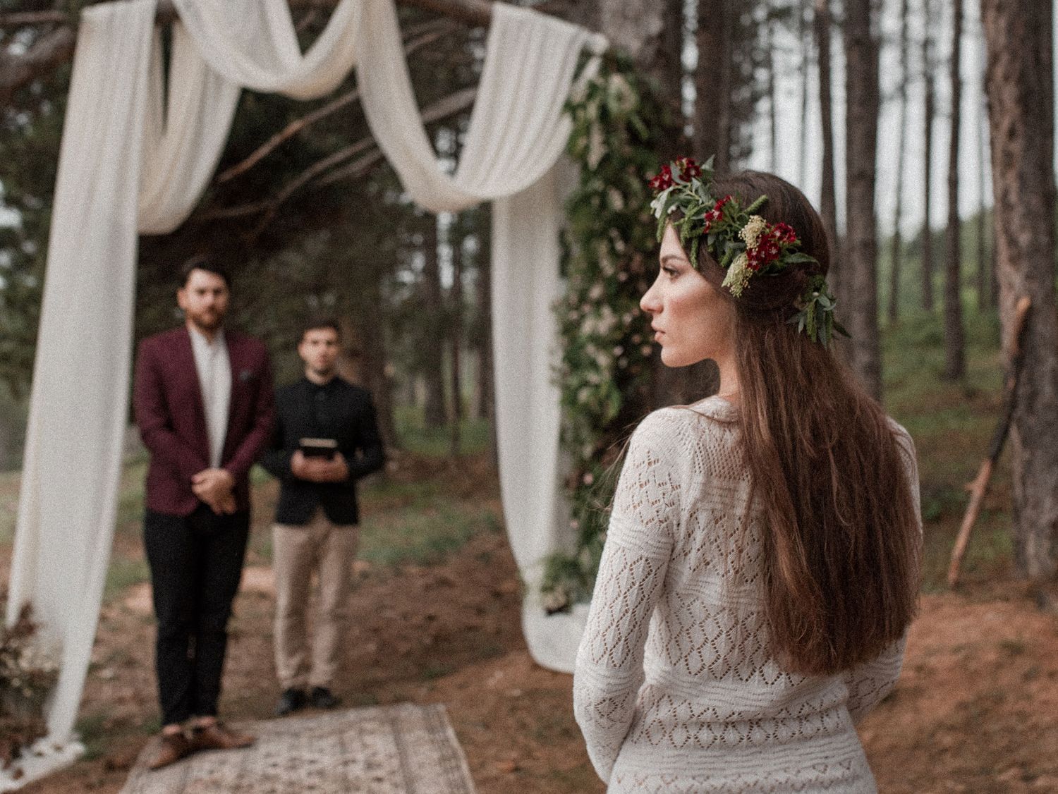 Outdoor forest wedding ceremony setup with white fabric arch and bride in lace dress wearing floral crown.