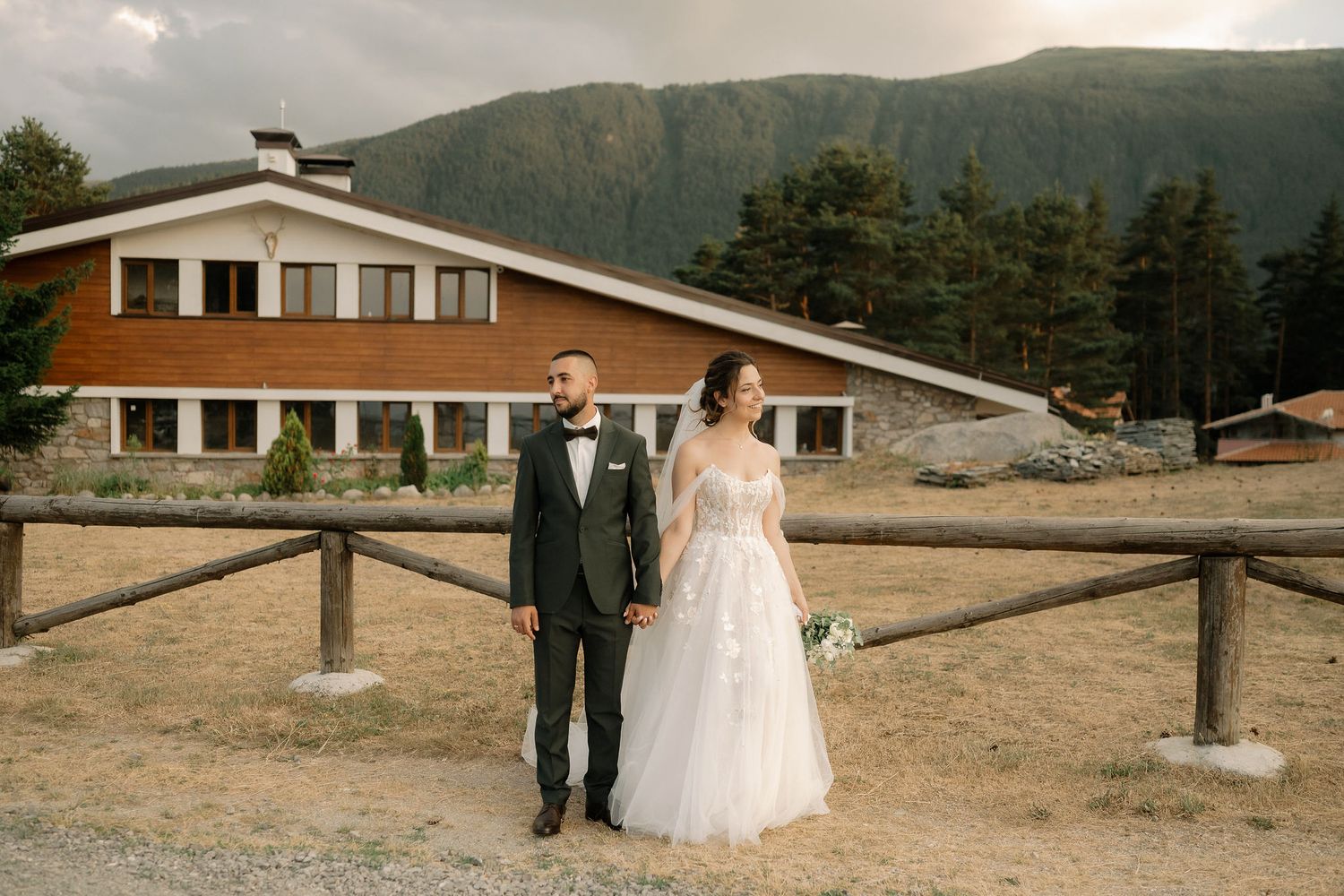 Wedding couple poses against mountain backdrop with rustic buildings and dramatic cloudy sky in panoramic view.