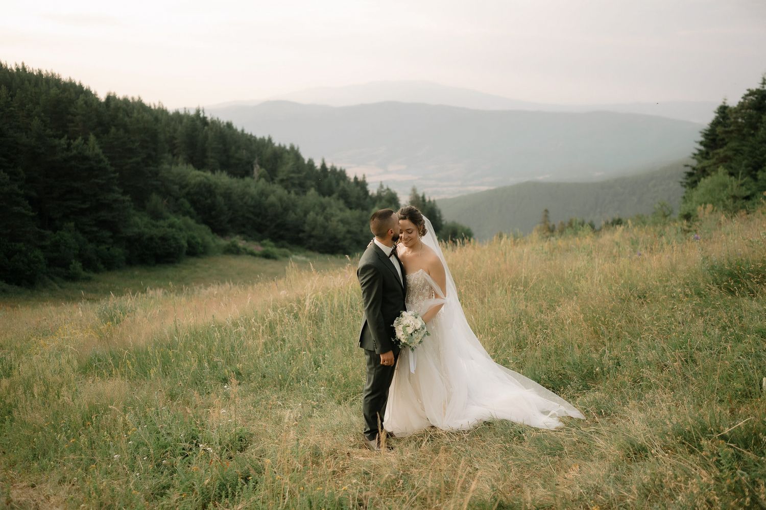 A couple in wedding attire embrace on a hilltop meadow overlooking misty mountain ranges at dusk.