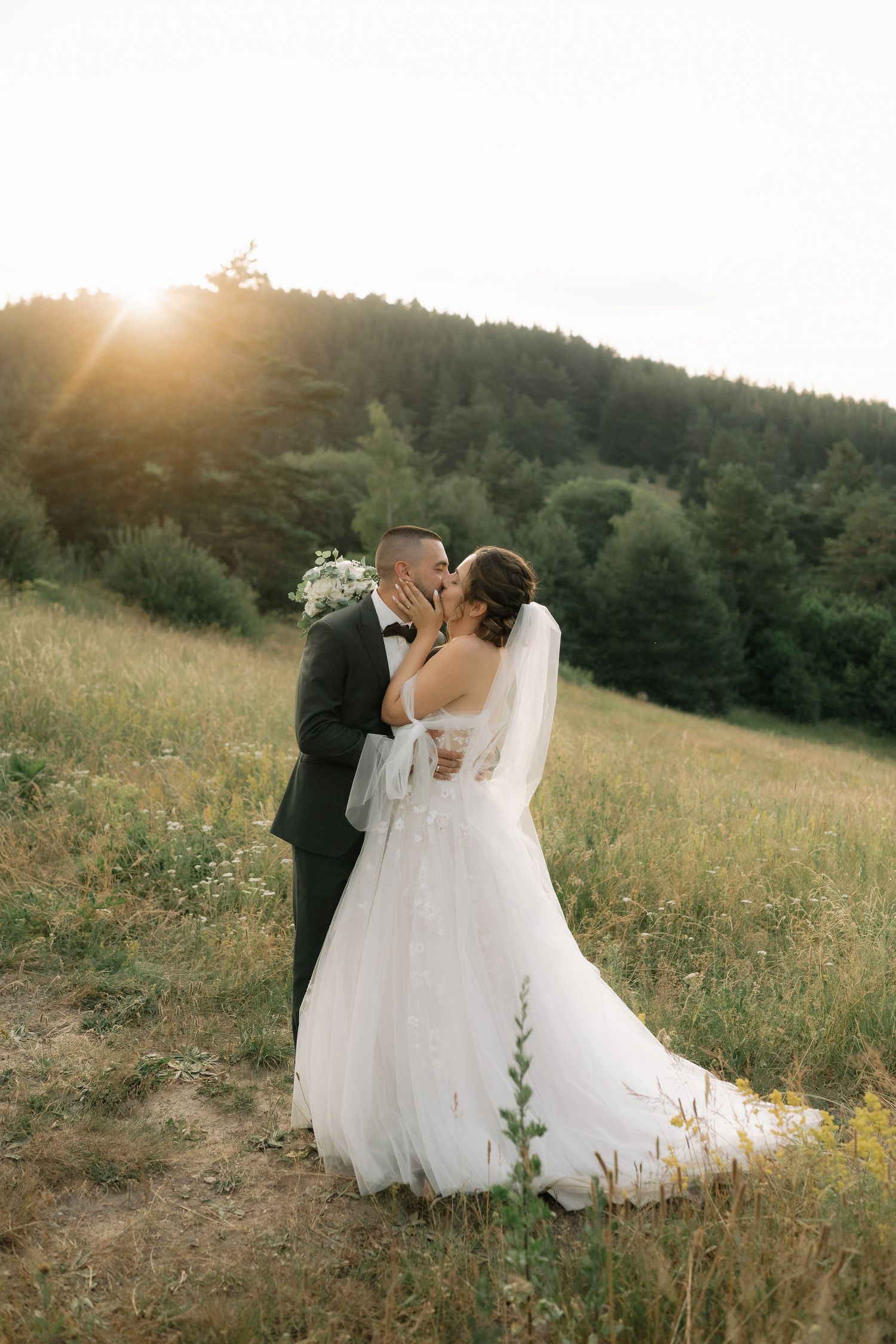 A romantic wedding couple embraces in a sunlit meadow with mountains and forest in the background during golden hour.