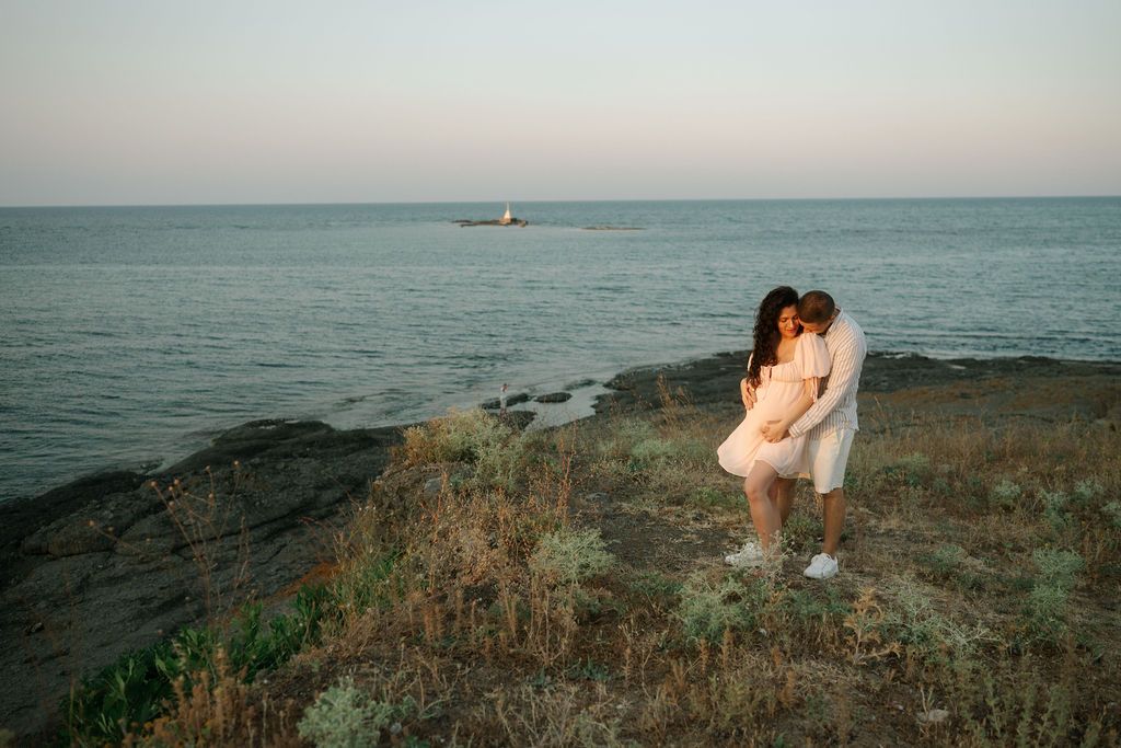 A romantic coastal photoshoot at sunset shows a couple embracing on rugged cliffs overlooking the ocean.
