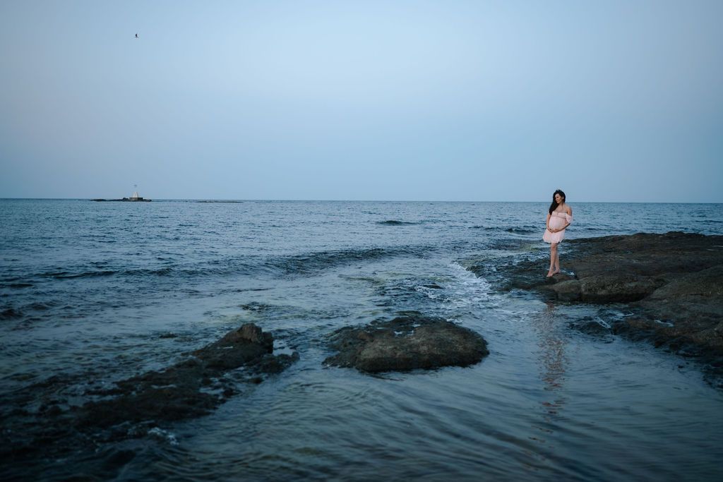 A person stands on a rocky outcrop jutting into the calm ocean during blue hour.