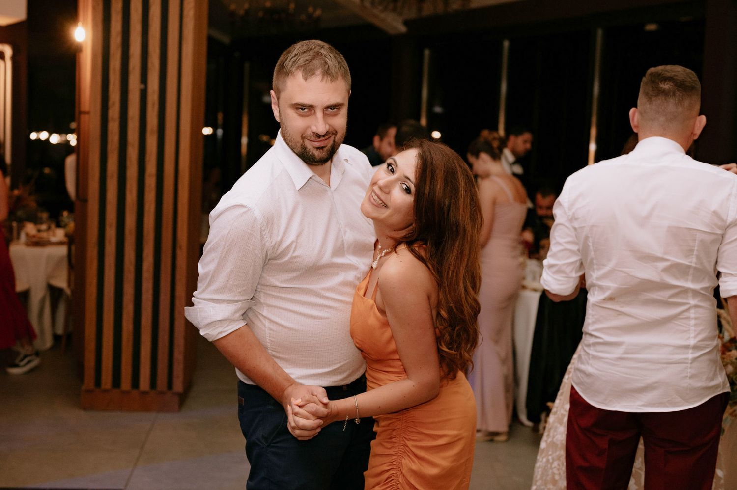 Couples dancing together at a social event wearing white shirts and orange dresses in a dimly lit venue.