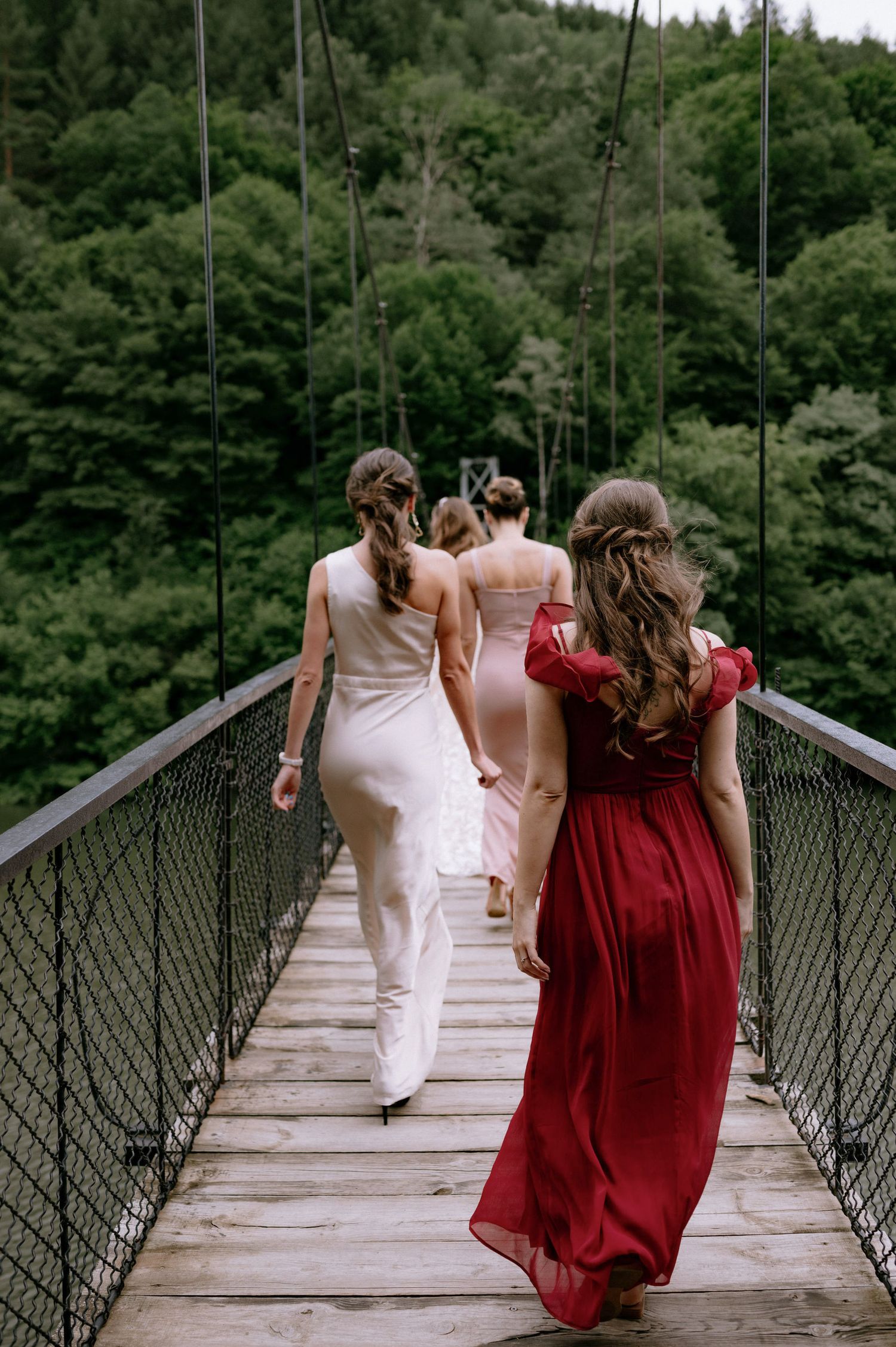 A group in white and burgundy dresses runs playfully across a suspended footbridge surrounded by lush forest.