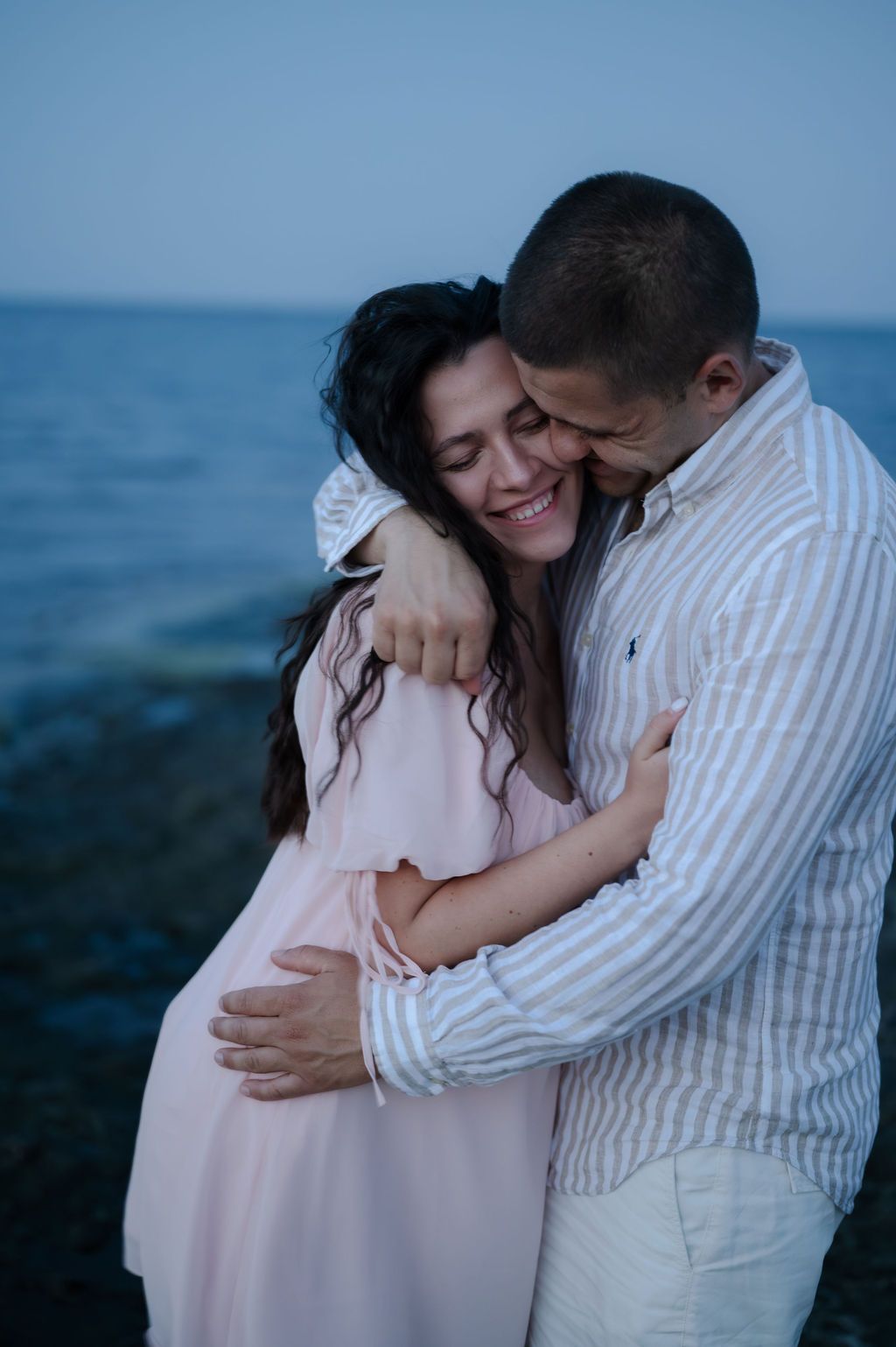 A couple embraces on the beach during a romantic sunset photoshoot wearing white and pink outfits.