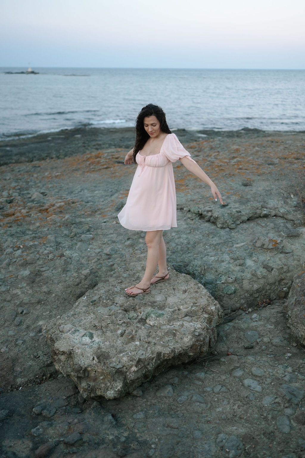 A person in a flowing pink dress balances gracefully on a rocky shoreline during sunset.