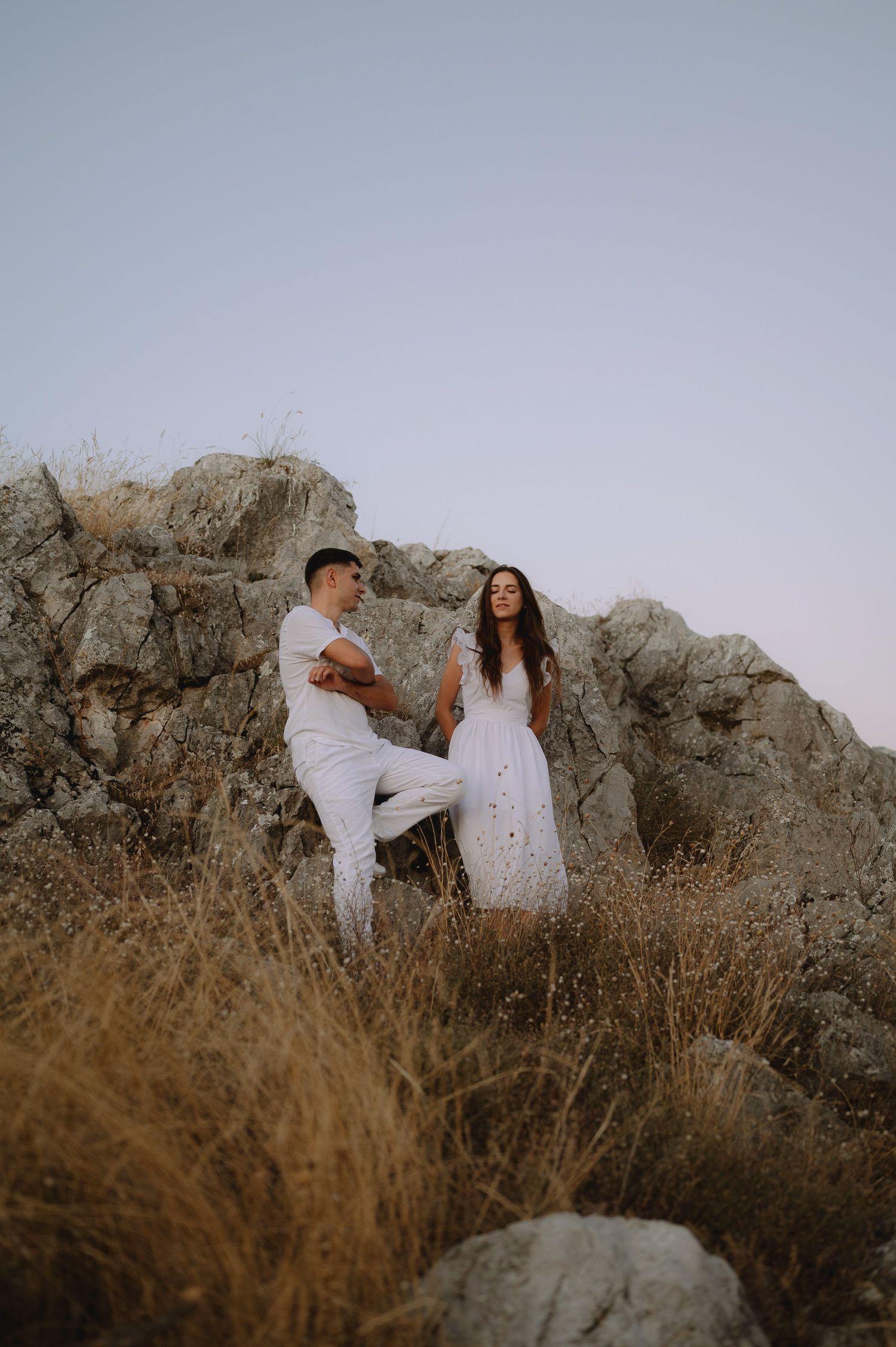 A couple in white outfits poses together against rocky terrain and dry grass at sunset for romantic photos.