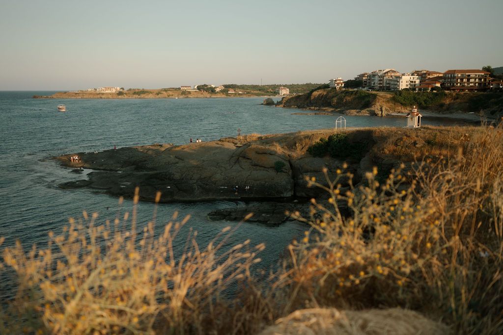Coastal landscape with golden grasses and rugged cliffs overlooking a serene blue ocean bay at sunset.