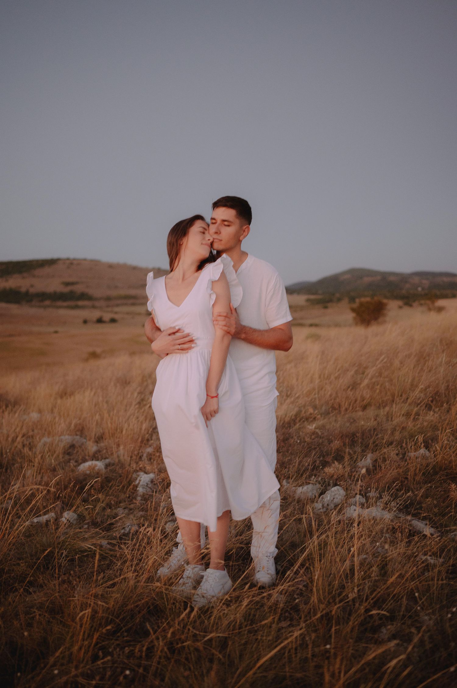 A romantic couple in white outfits embrace in a golden field at sunset with mountains in the background.