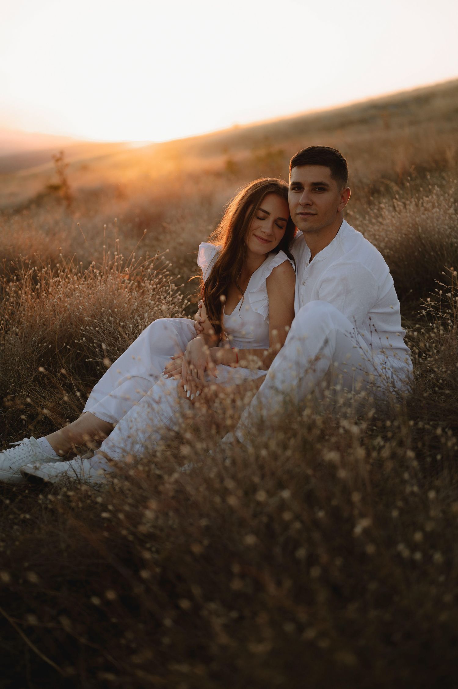 Romantic couple in white outfits embracing in a field during golden hour sunset.