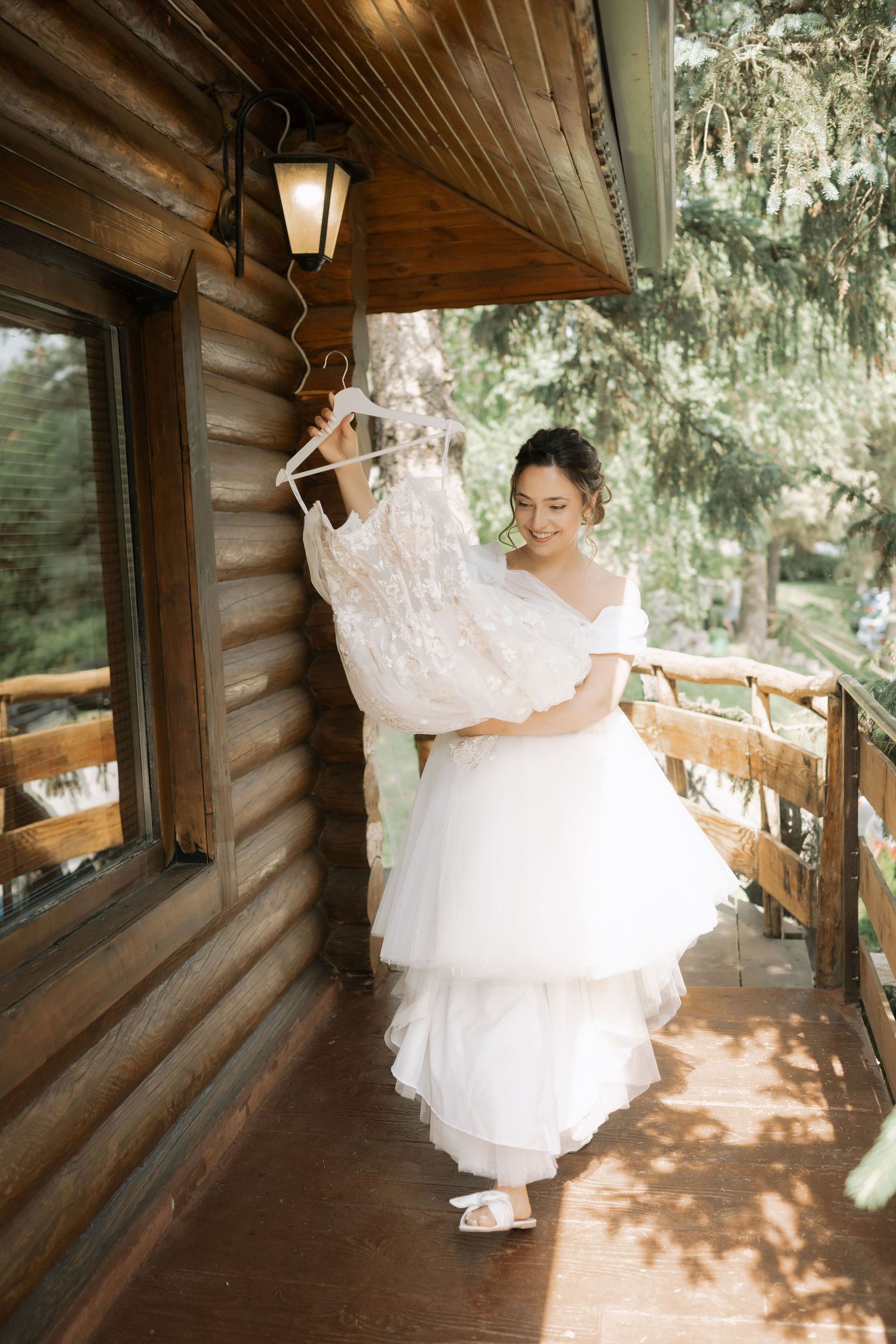 A bride in a flowing white wedding dress twirls playfully on a rustic wooden cabin porch in dappled sunlight.