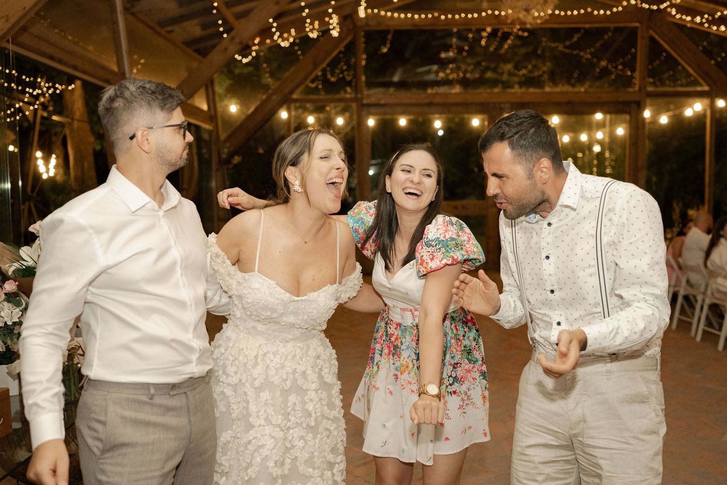 Wedding guests share joyful laughter together on the dance floor under warm string lights at an indoor reception.