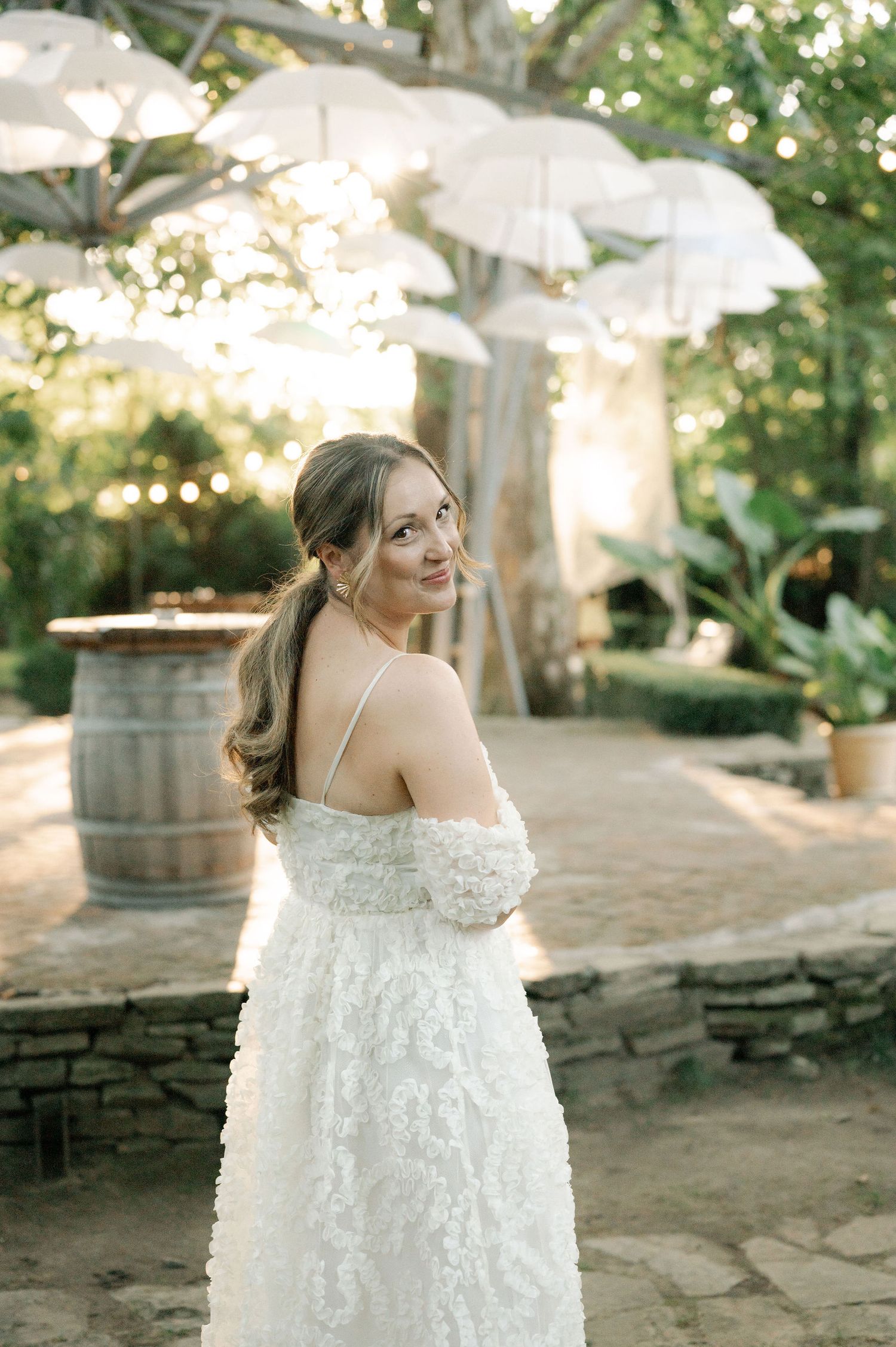 A white lace wedding dress is showcased in a series of outdoor photos with stone walls and greenery in the background.