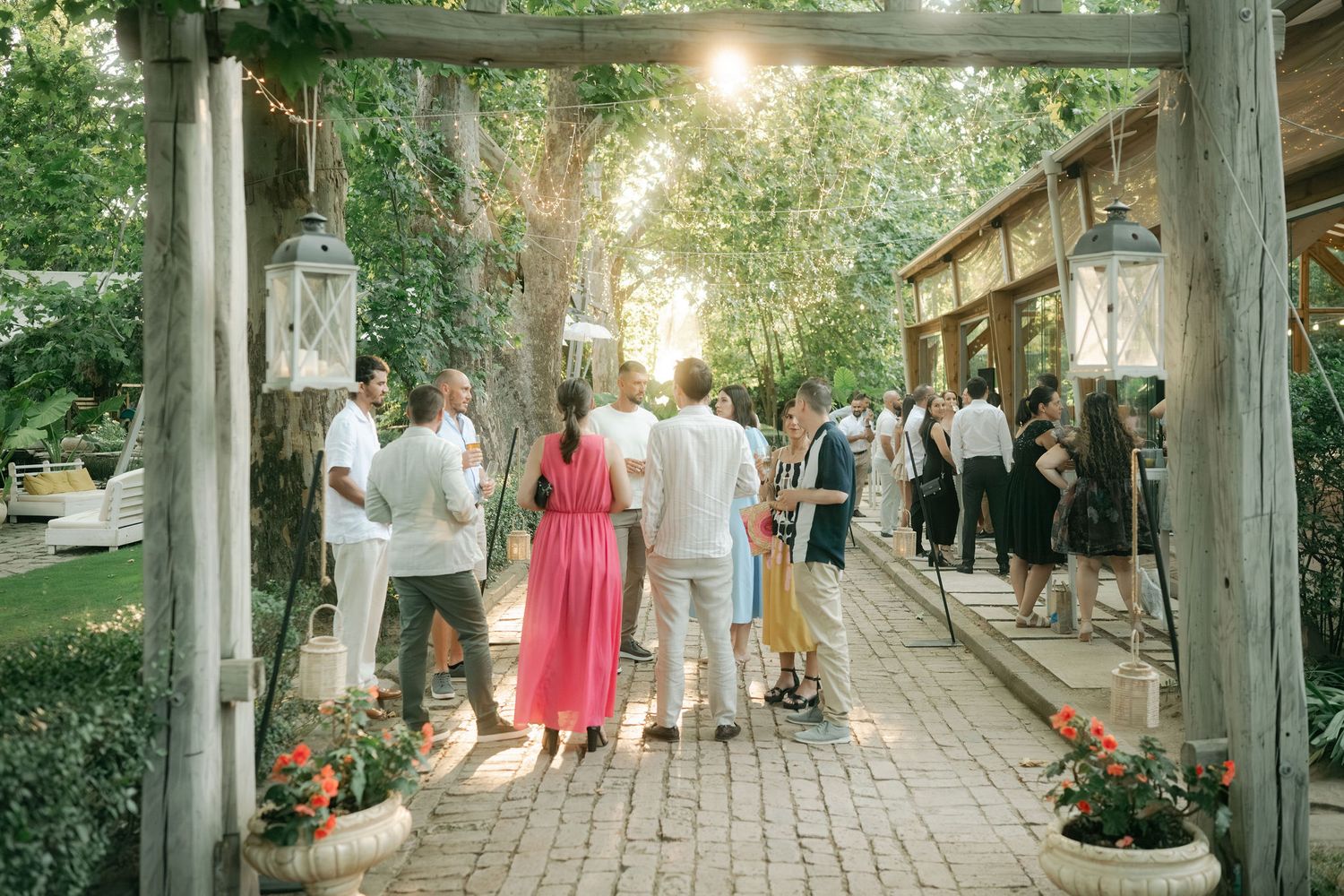 Guests walk along a rustic wooden boardwalk surrounded by lush greenery and string lights at an outdoor event.