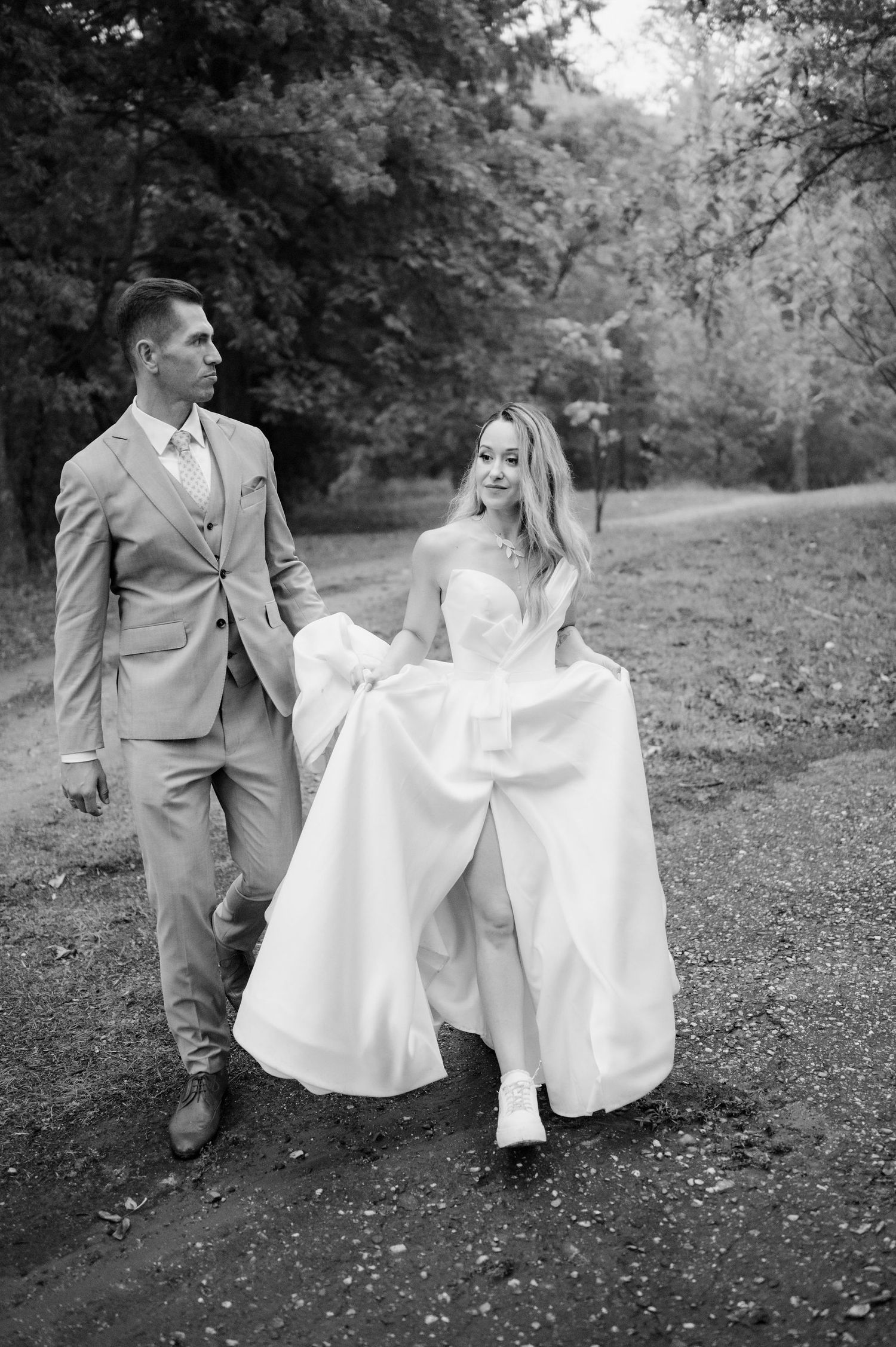 A couple in wedding attire walking together on a path through nature, captured in black and white.